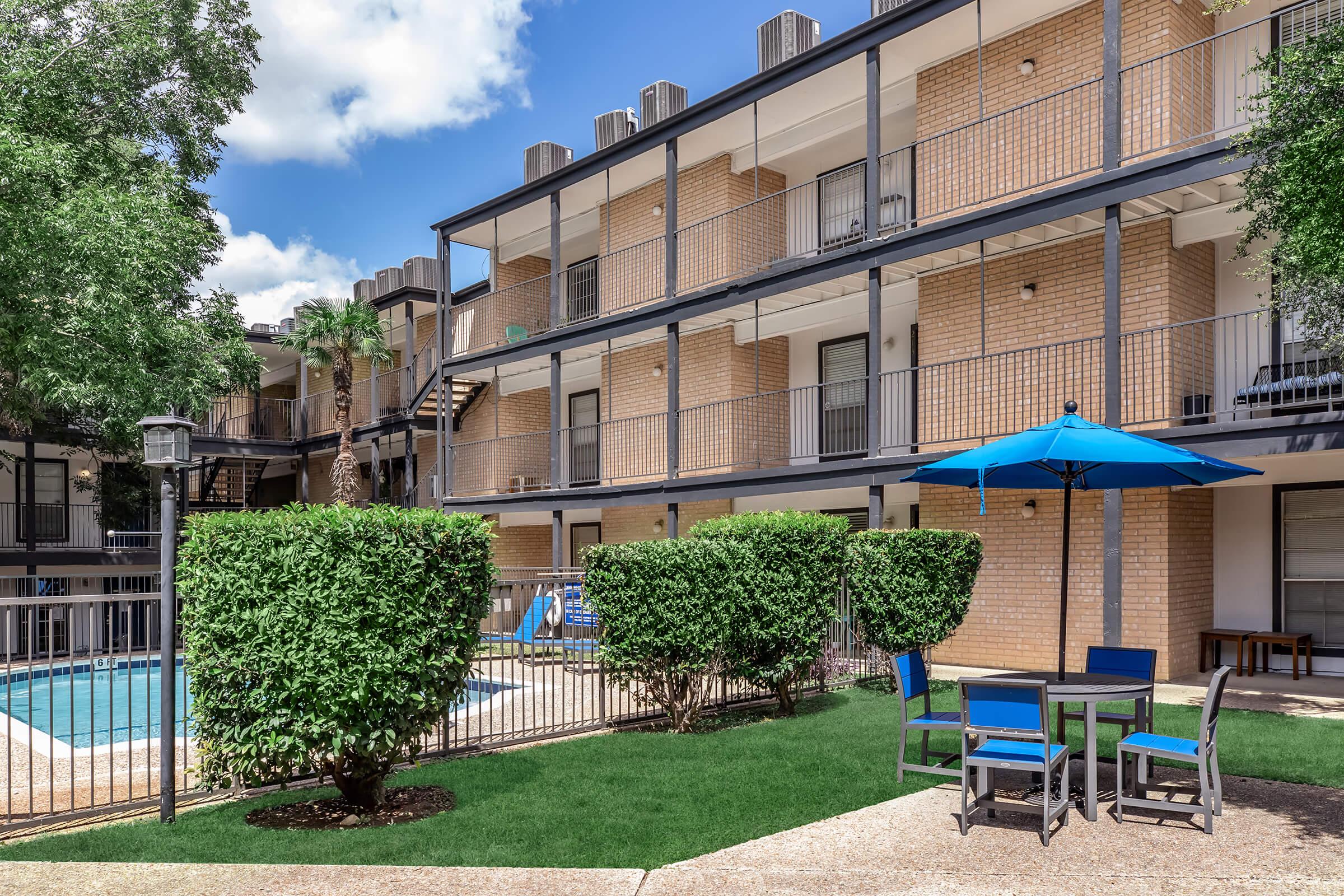 A view of a multi-story apartment complex featuring a swimming pool in the center, surrounded by well-maintained green shrubs and a small seating area with blue chairs and an umbrella. The building has balconies and is set against a blue sky with scattered clouds.