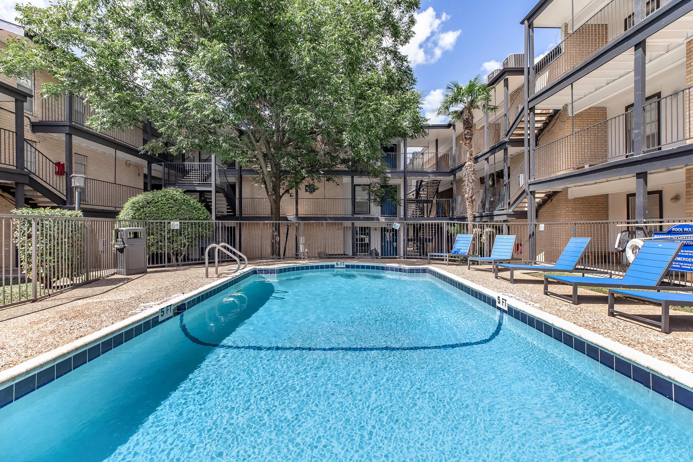 A clear swimming pool surrounded by lounge chairs, set within a well-maintained courtyard of an apartment complex. Lush green trees provide shade, and the buildings feature multiple balconies and staircases. The sky is bright with a few clouds, creating a welcoming and relaxed atmosphere.
