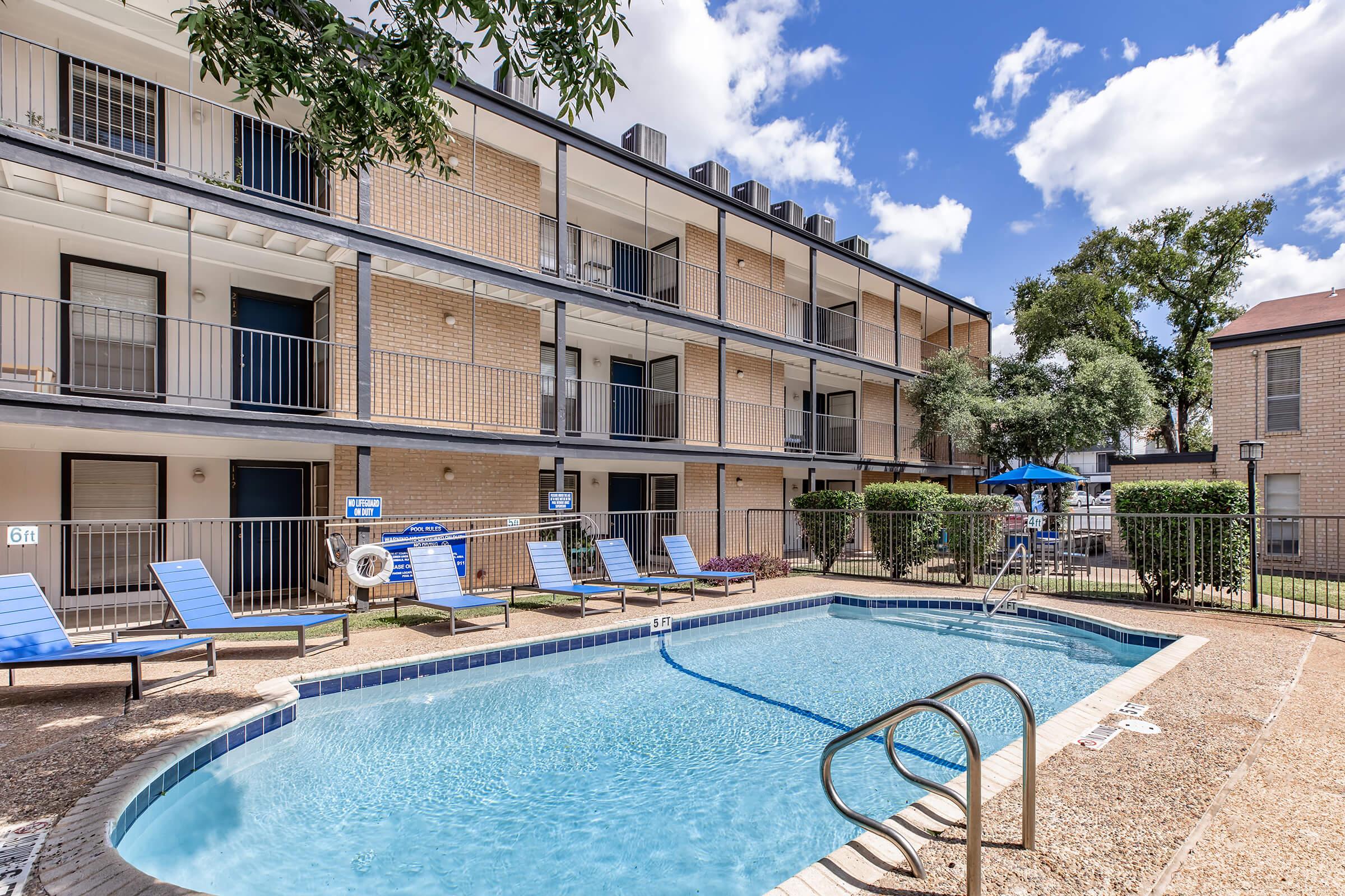 A photo of an apartment complex featuring a small pool area. The pool is surrounded by lounge chairs and has a fence for safety. The building has three levels with balconies, and there are trees and shrubs around the pool area. The sky is partly cloudy, creating a bright atmosphere.