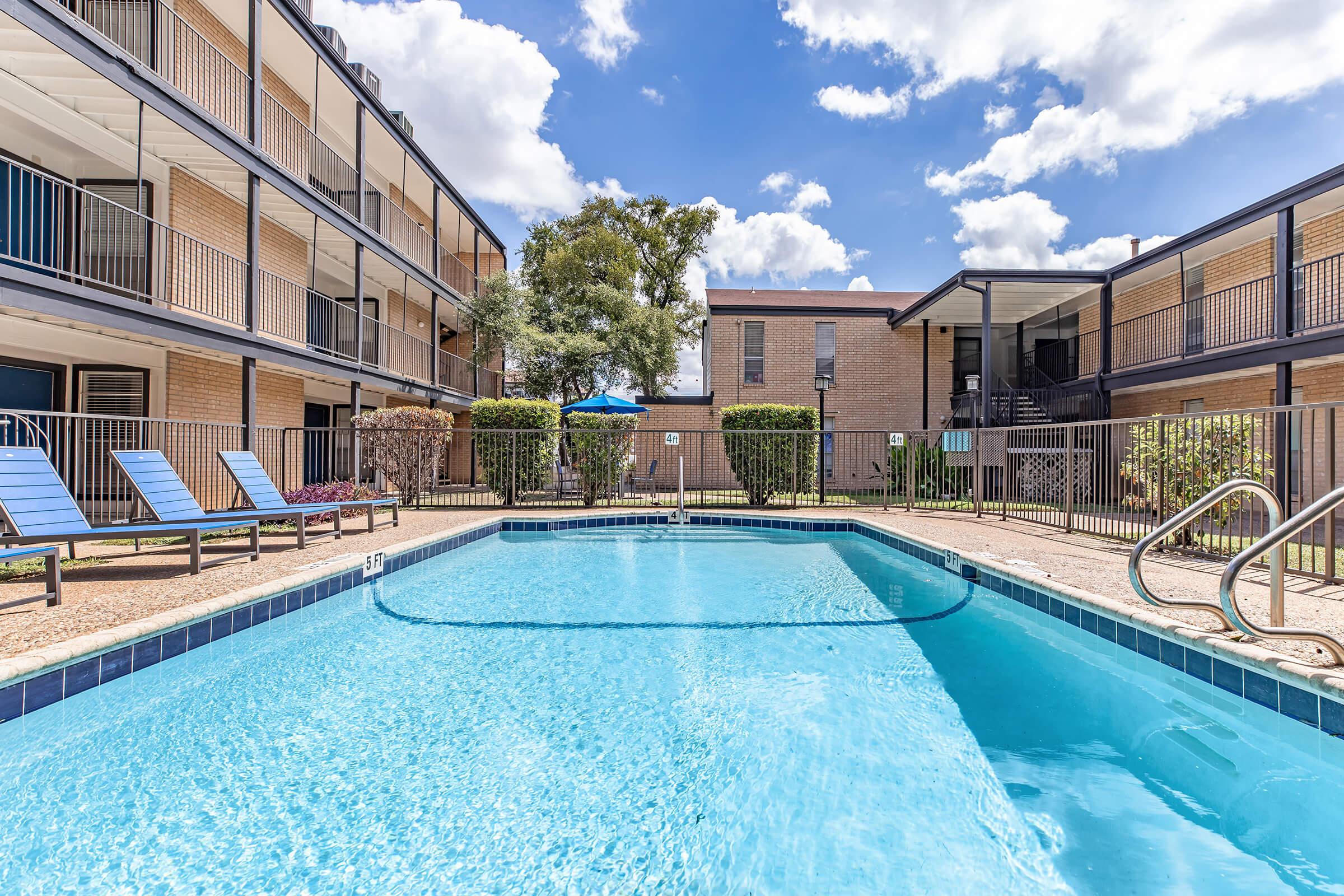 A clear blue swimming pool surrounded by colorful lounge chairs, with two-story buildings in the background. Green shrubs and trees provide a refreshing atmosphere, and a bright sky with fluffy white clouds adds to the inviting scene.