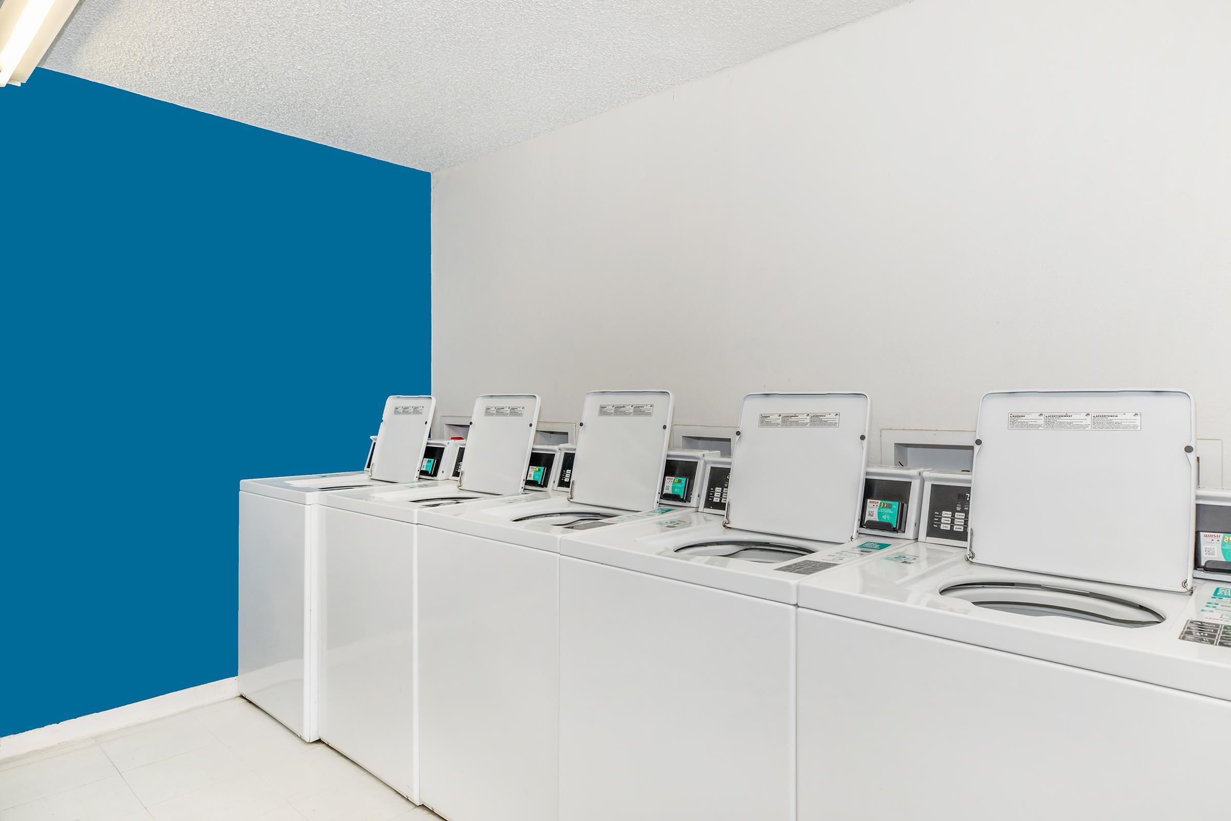 A row of white washing machines in a laundry room, featuring a blue accent wall on one side. The machines are front-loading with lids open, and the floor is tiled. The overall atmosphere is clean and well-lit.