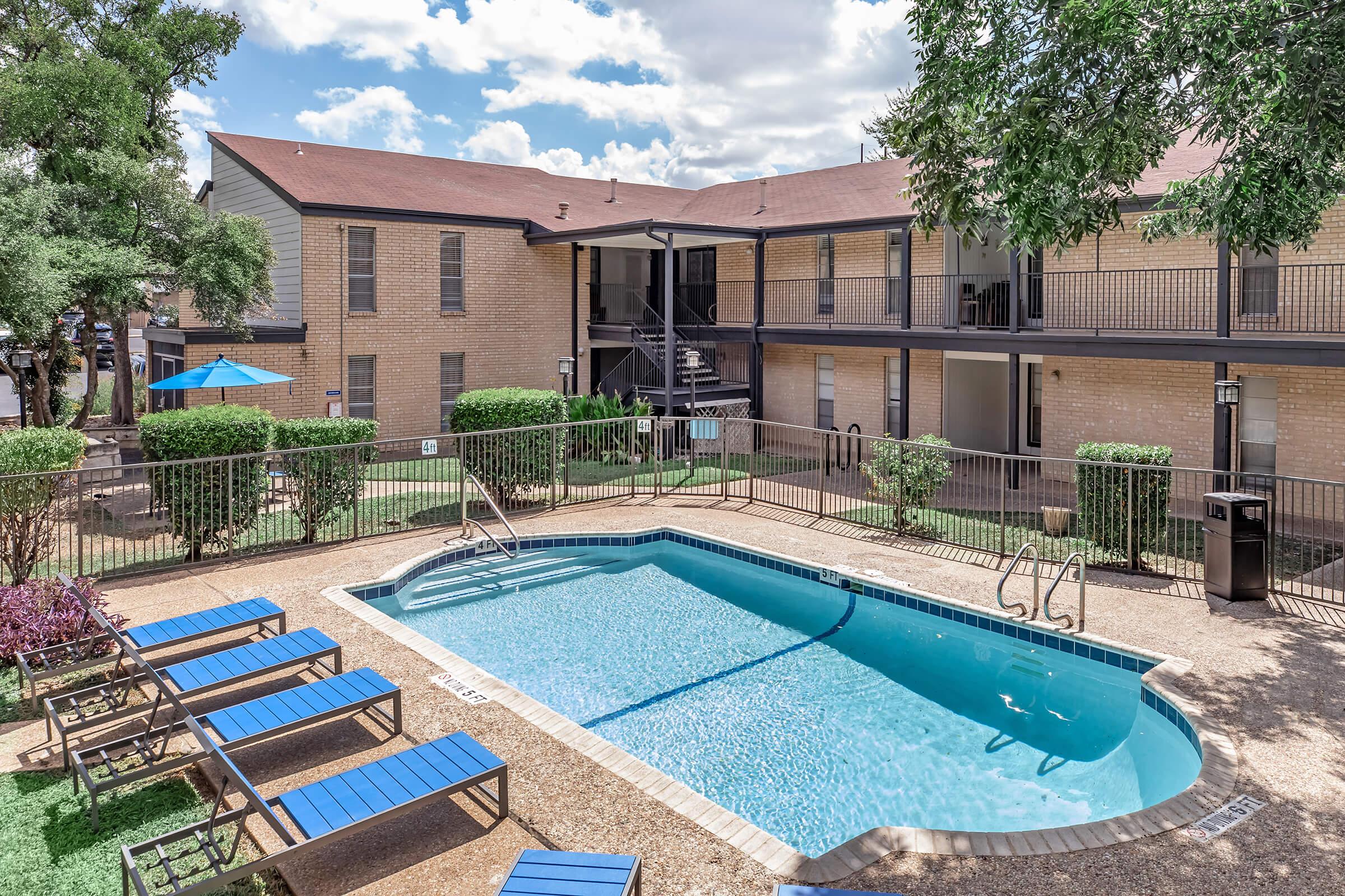 A clean, inviting outdoor pool area surrounded by blue lounge chairs and well-maintained greenery. The pool is nestled between two buildings, with a staircase leading to upper-level apartments. Bright blue sky with clouds adds a cheerful ambiance to the scene.