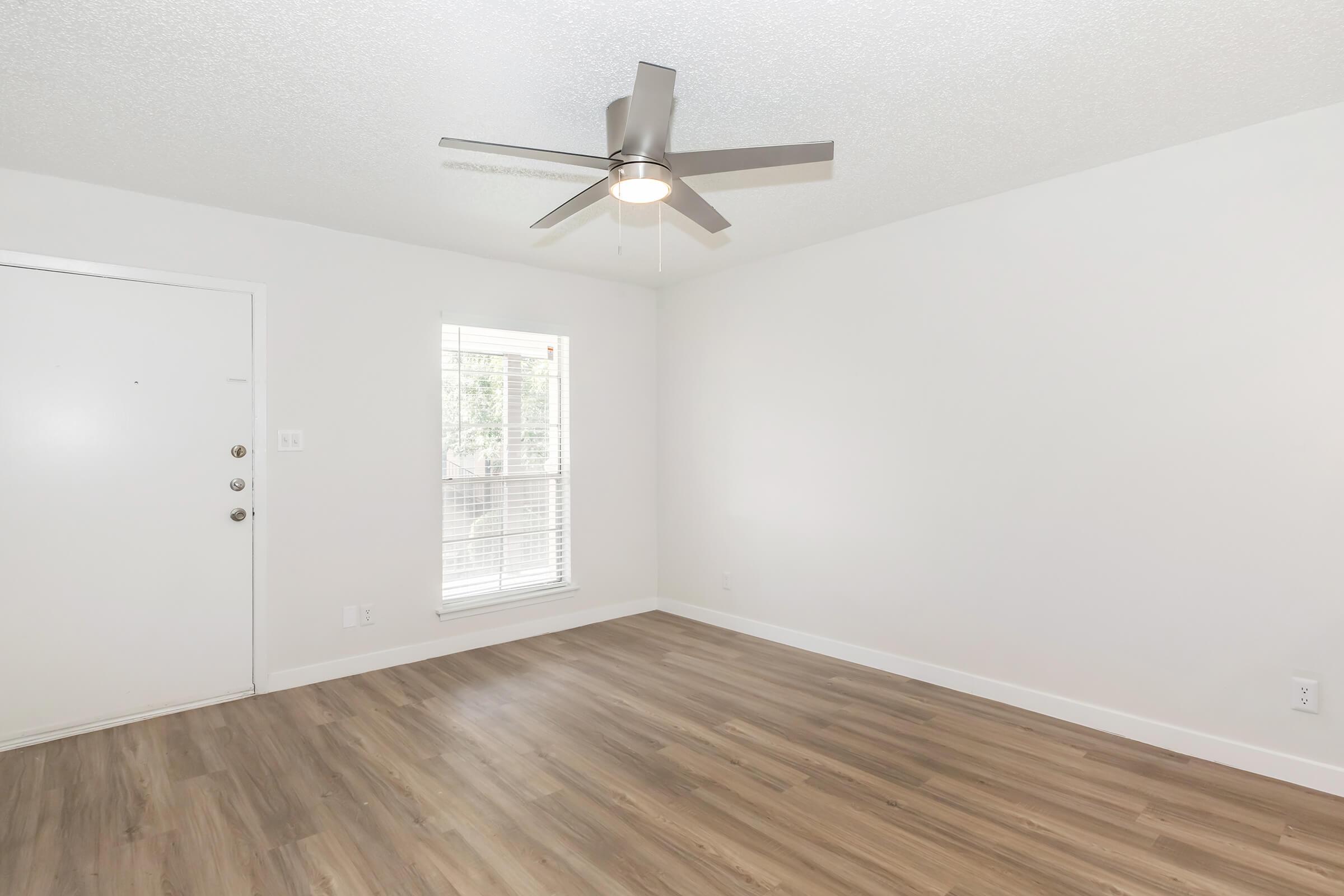 Interior view of a well-lit room with a light-colored wall and wooden flooring. A ceiling fan is mounted in the center. There's a white door on the left and a window on the right, allowing natural light to fill the space. The overall appearance is clean and modern.