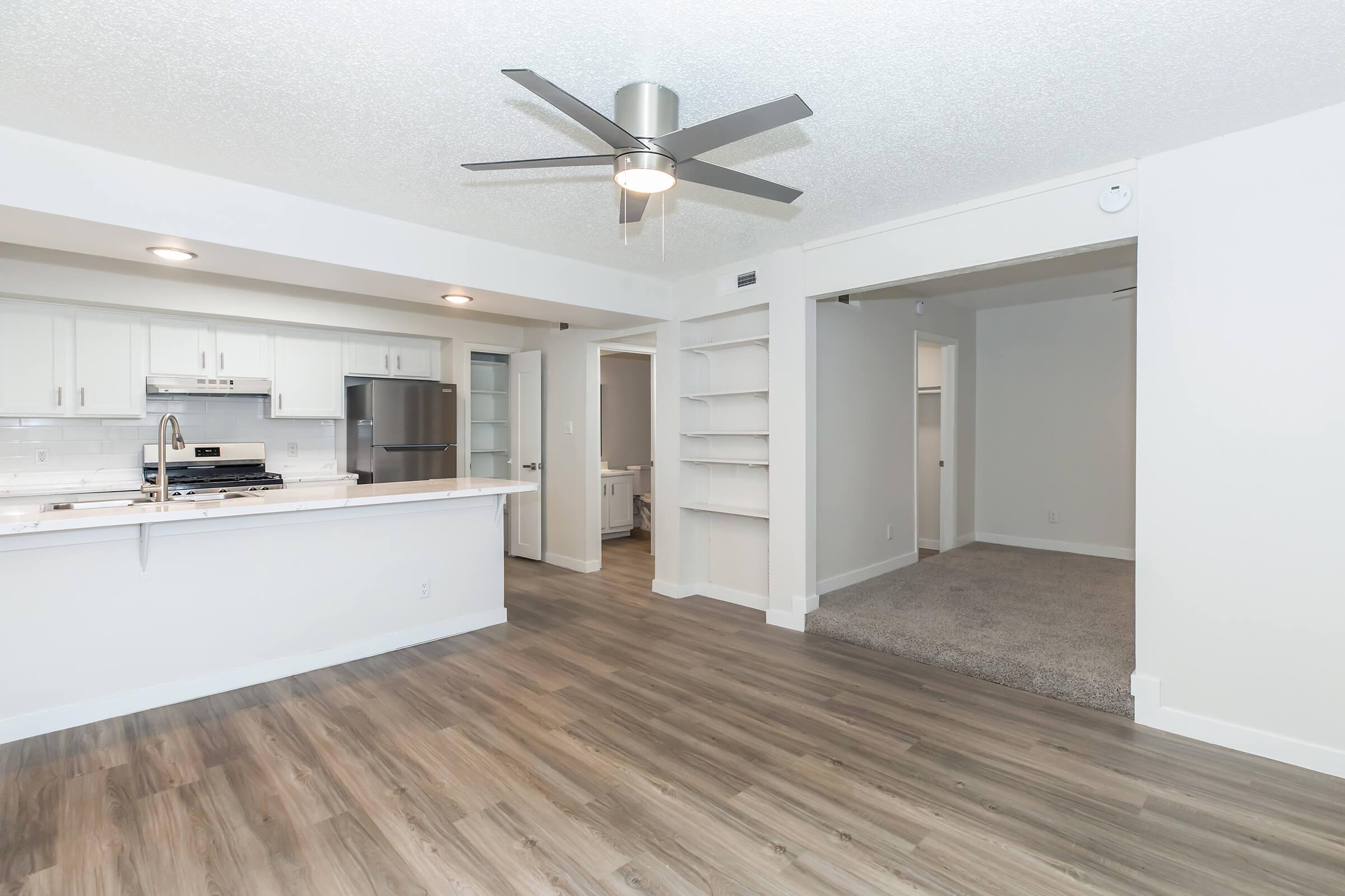 A spacious, modern living area with light-colored walls and wood plank flooring. It features an open concept design connecting the kitchen and living room, with stainless steel appliances and a ceiling fan. A carpeted area is visible leading to another room, along with built-in shelves.
