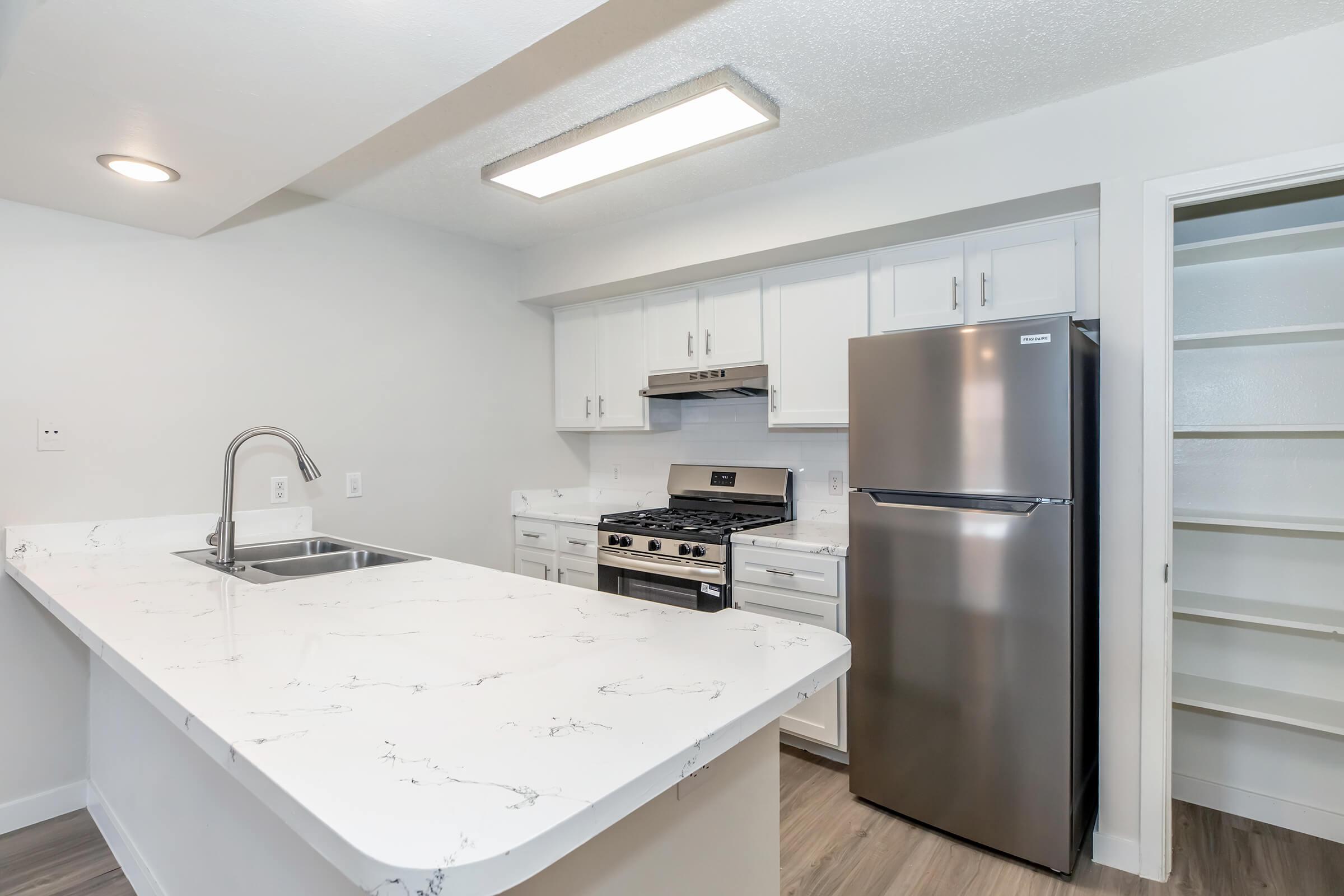 Bright, modern kitchen featuring white cabinetry, stainless steel appliances, and a large island with a marble countertop. A gas stove is positioned under a range hood, while a sleek refrigerator stands nearby. The space has a light, airy feel with ample storage and open shelving.