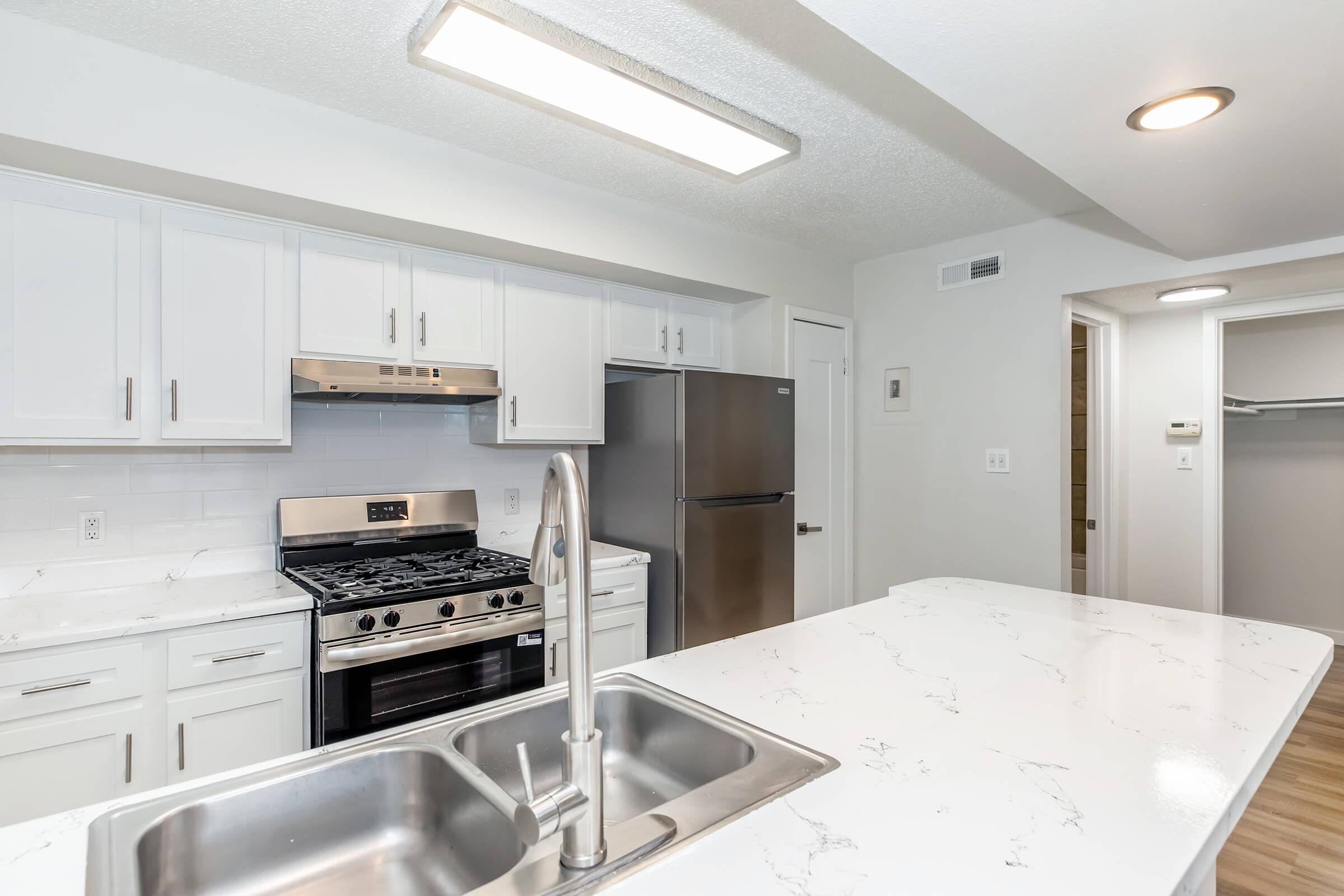 Modern kitchen featuring white cabinetry, a stainless steel gas stove, and an overhead exhaust hood. A large island with a double sink is in the foreground, and a refrigerator is visible in the background. The space has neutral-colored walls and a well-lit atmosphere.