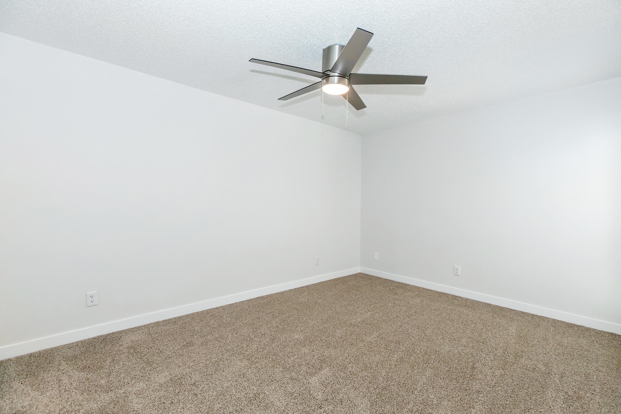 A spacious empty room with a light-colored carpet and white walls. The ceiling features a modern silver ceiling fan. There are no furnishings, creating a bright and airy atmosphere. The room has a clean, minimalist design suitable for various uses.