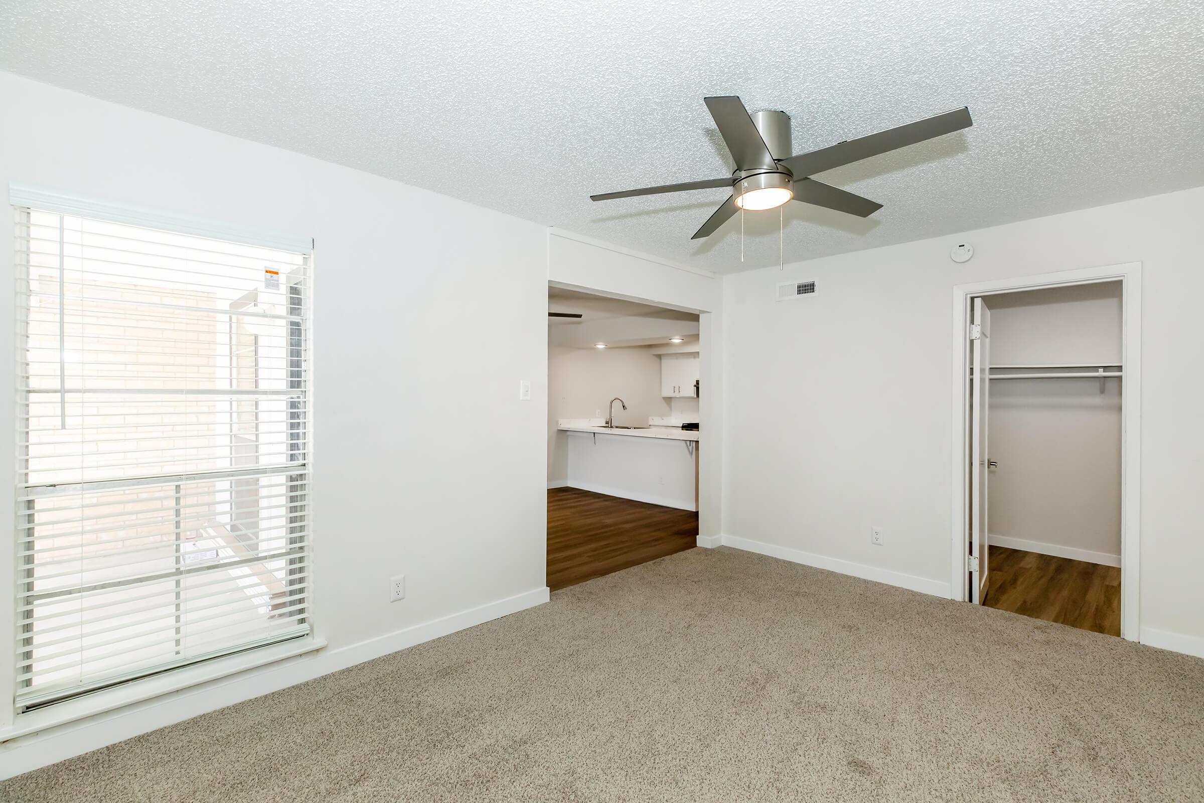 A spacious, carpeted living area featuring a ceiling fan, a window with blinds, and an open view towards a kitchen. There is a closet with a door on the right and a light-colored wall, creating a bright and airy atmosphere in a modern apartment setting.