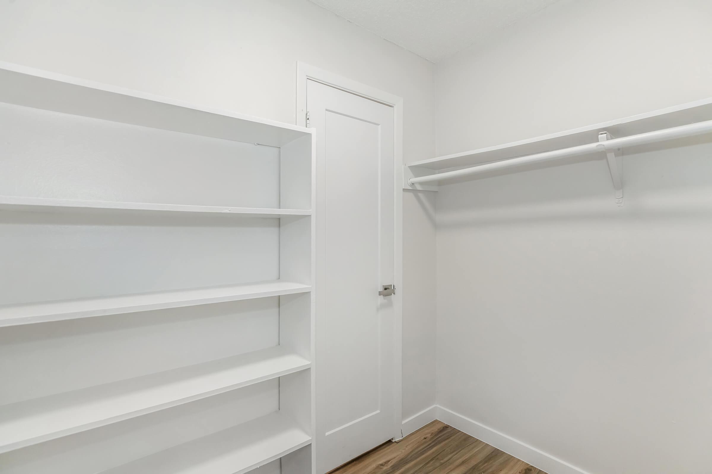 Empty room with white walls and a light wood floor featuring built-in white shelves on the left and a single shelf mounted on the right wall. A closed door is visible in the background, contributing to a minimalist and organized appearance. Ideal for storage or as a closet space.
