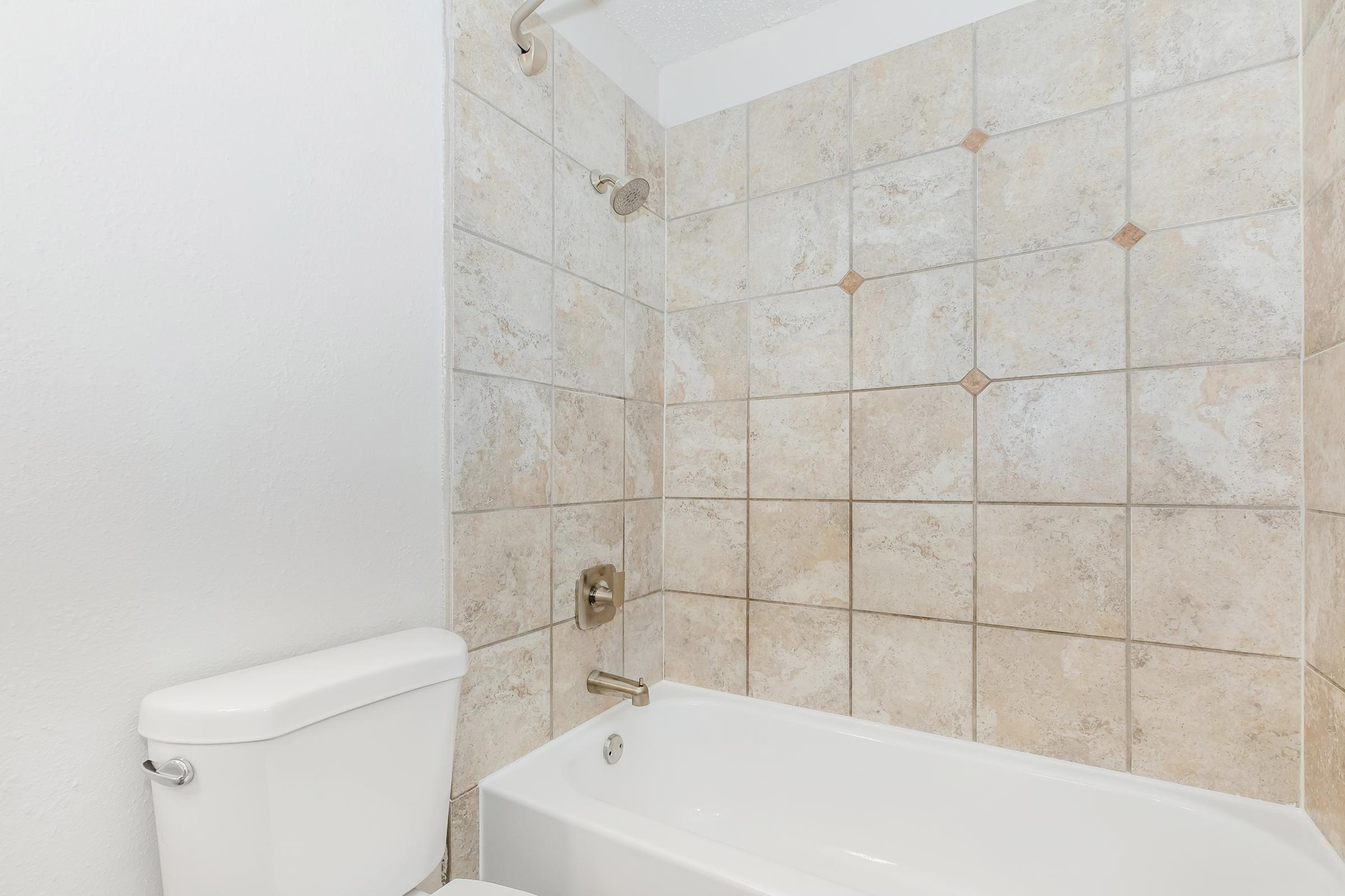 A modern bathroom featuring a clean bathtub and a wall-mounted showerhead. The walls are decorated with light-colored tiles, while the toilet is visible on the left side of the image. The overall design is simple and contemporary, with a neutral color palette.