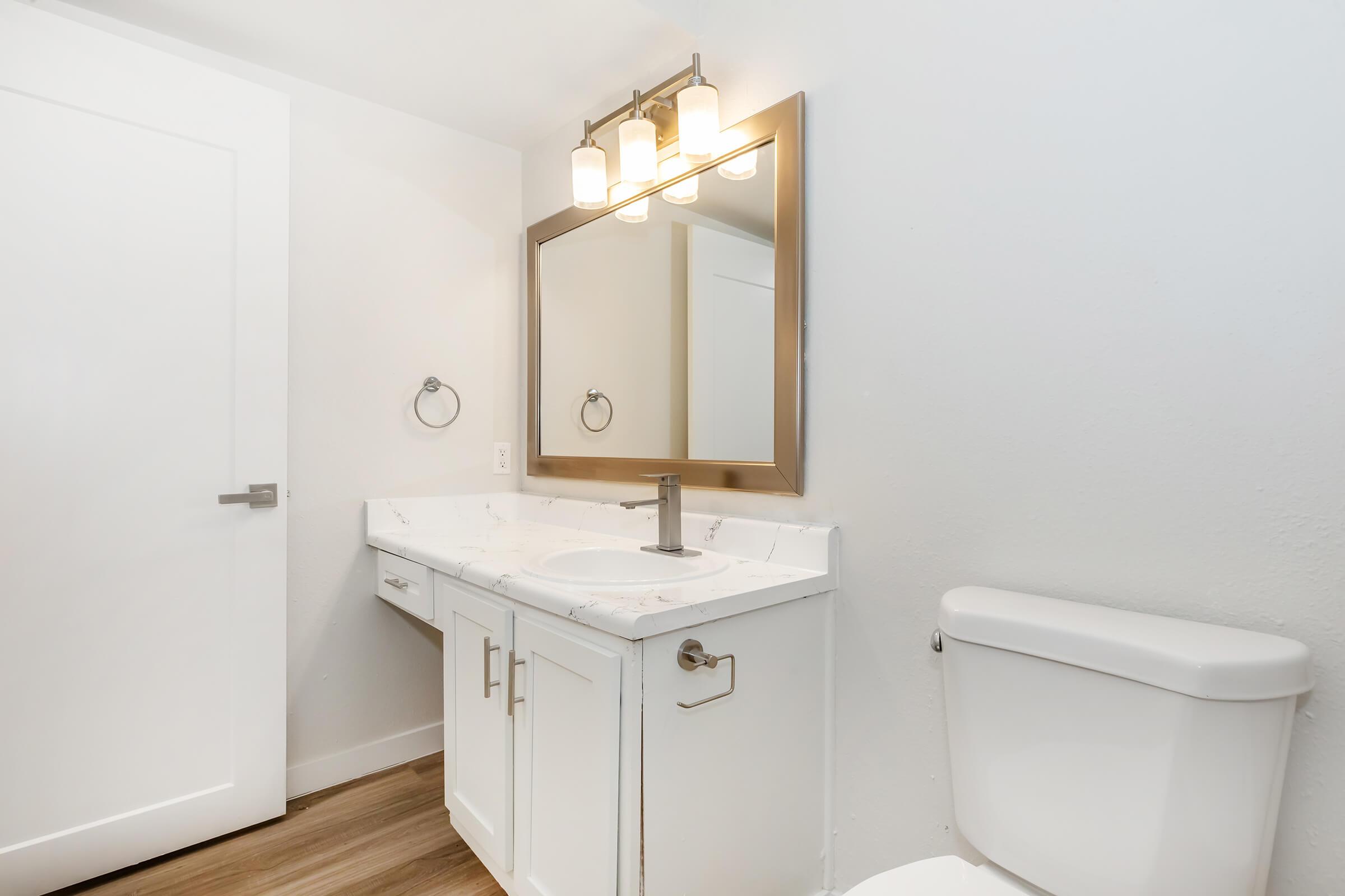 A modern bathroom featuring a white vanity with a marble countertop, a large framed mirror above the sink, a wall-mounted towel holder, and a sleek toilet. The walls are painted white, and the flooring is a light wood tone, creating a bright and clean aesthetic.