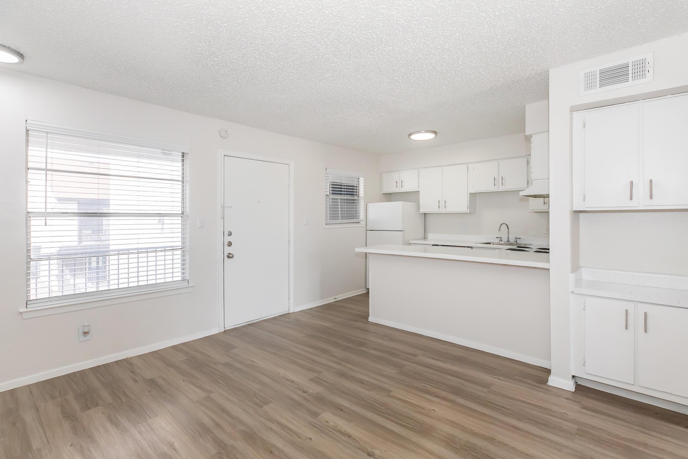 Bright and modern kitchen and living area featuring white cabinets, a refrigerator, and a window with blinds. Light wood flooring adds warmth to the space. A door leads outside, and natural light fills the room. The layout is open and inviting, creating a comfortable atmosphere.