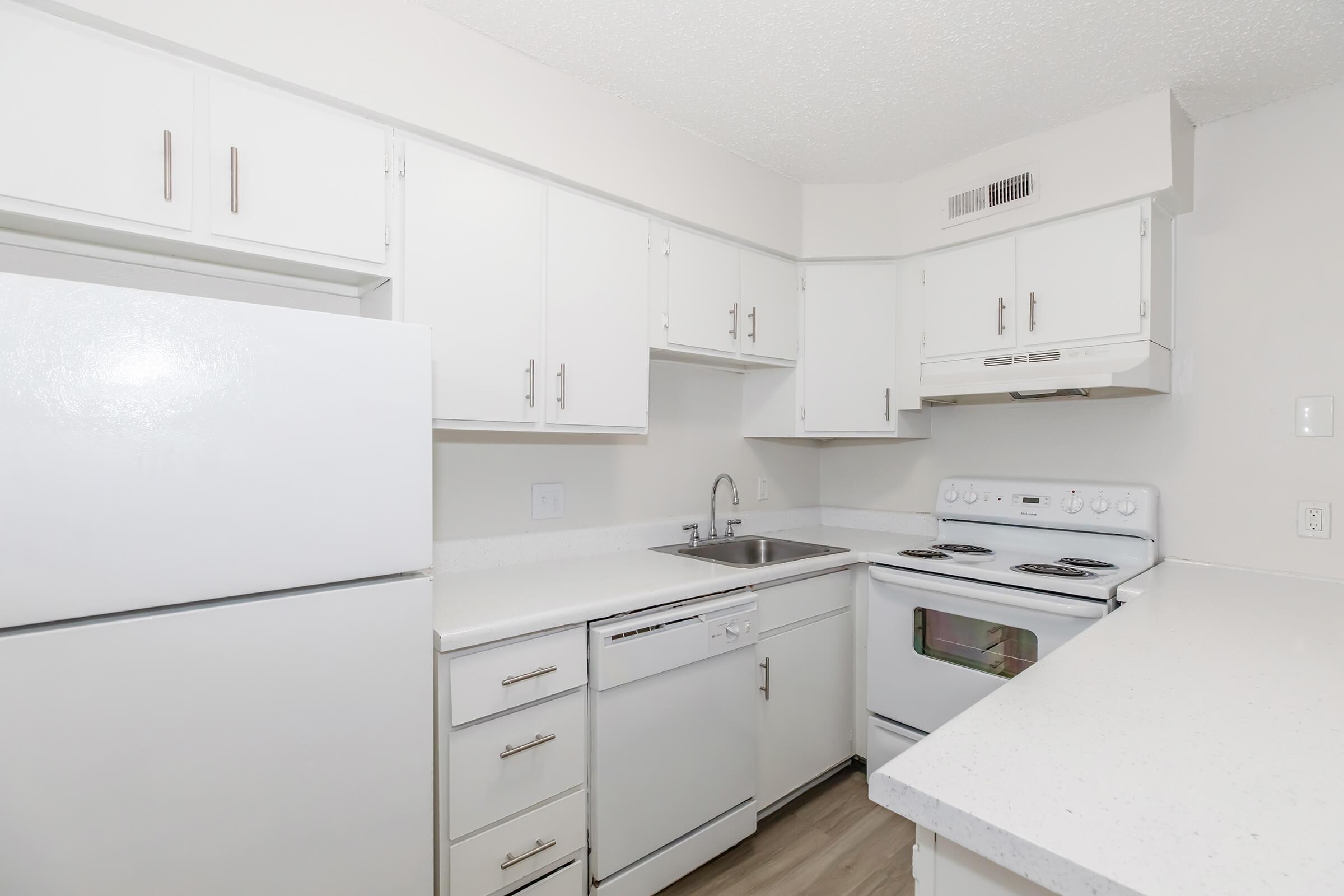 A modern kitchen featuring white cabinetry, a stainless steel sink, a white refrigerator, and a built-in dishwasher. The kitchen also includes a white stove and oven, with a light countertop and light wood-like flooring, creating a clean and bright appearance.