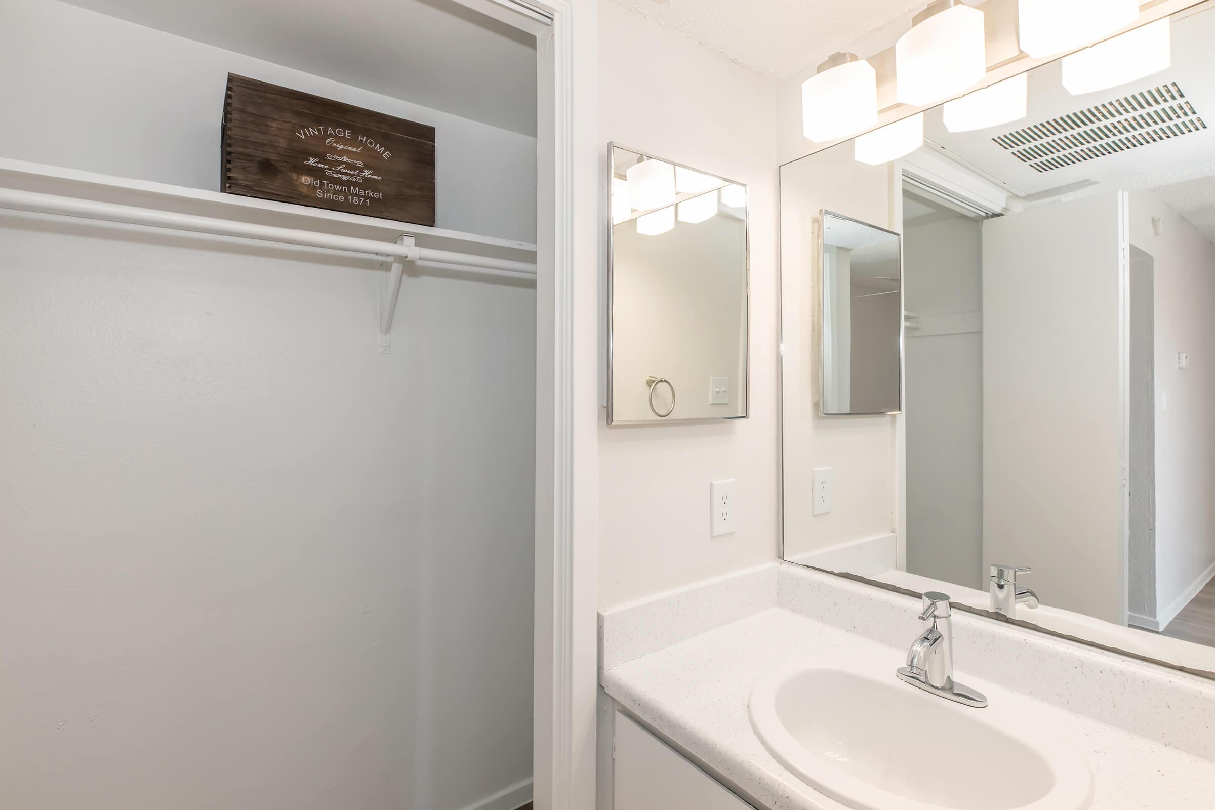 A clean, modern bathroom featuring a mirror above a white sink with a chrome faucet, a light fixture above, and a beige wall. To the left, there is a closet with a wooden sign labeled “VINTAGE HOME.” The space is well-lit and minimalistic, showcasing a neutral color palette.