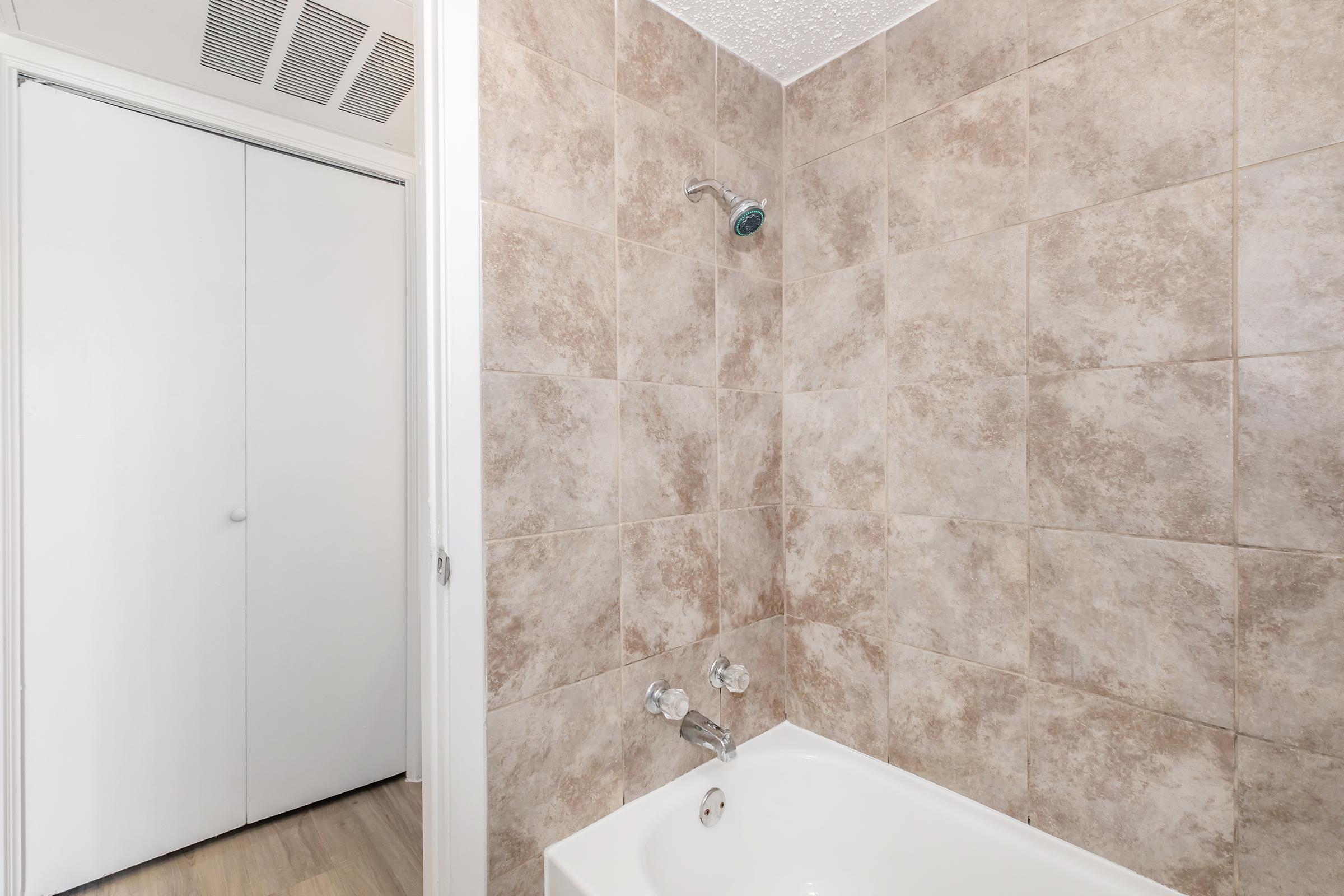 A bathroom showcasing a tiled shower area with a showerhead and a bathtub. The walls feature light brown tiles, and there is a white closet door partially visible in the background, complementing the neutral color scheme. The overall setting is clean and well-maintained.