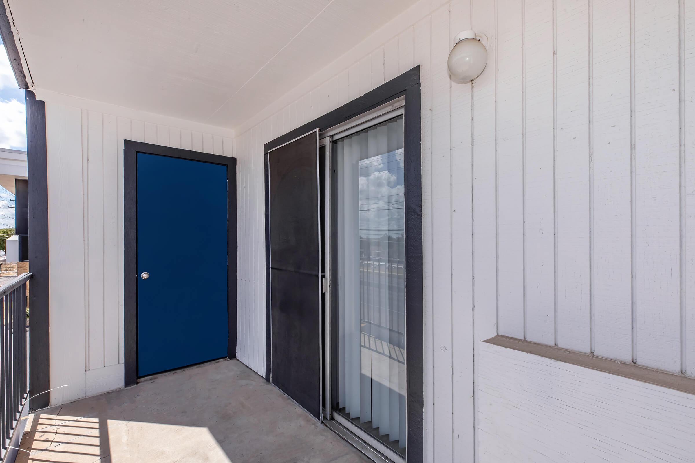 A porch area featuring a blue door and a sliding glass door. The wall is white wooden paneling, and there is a light fixture on the side. The floor is concrete, and the area is well-lit with natural light coming in.