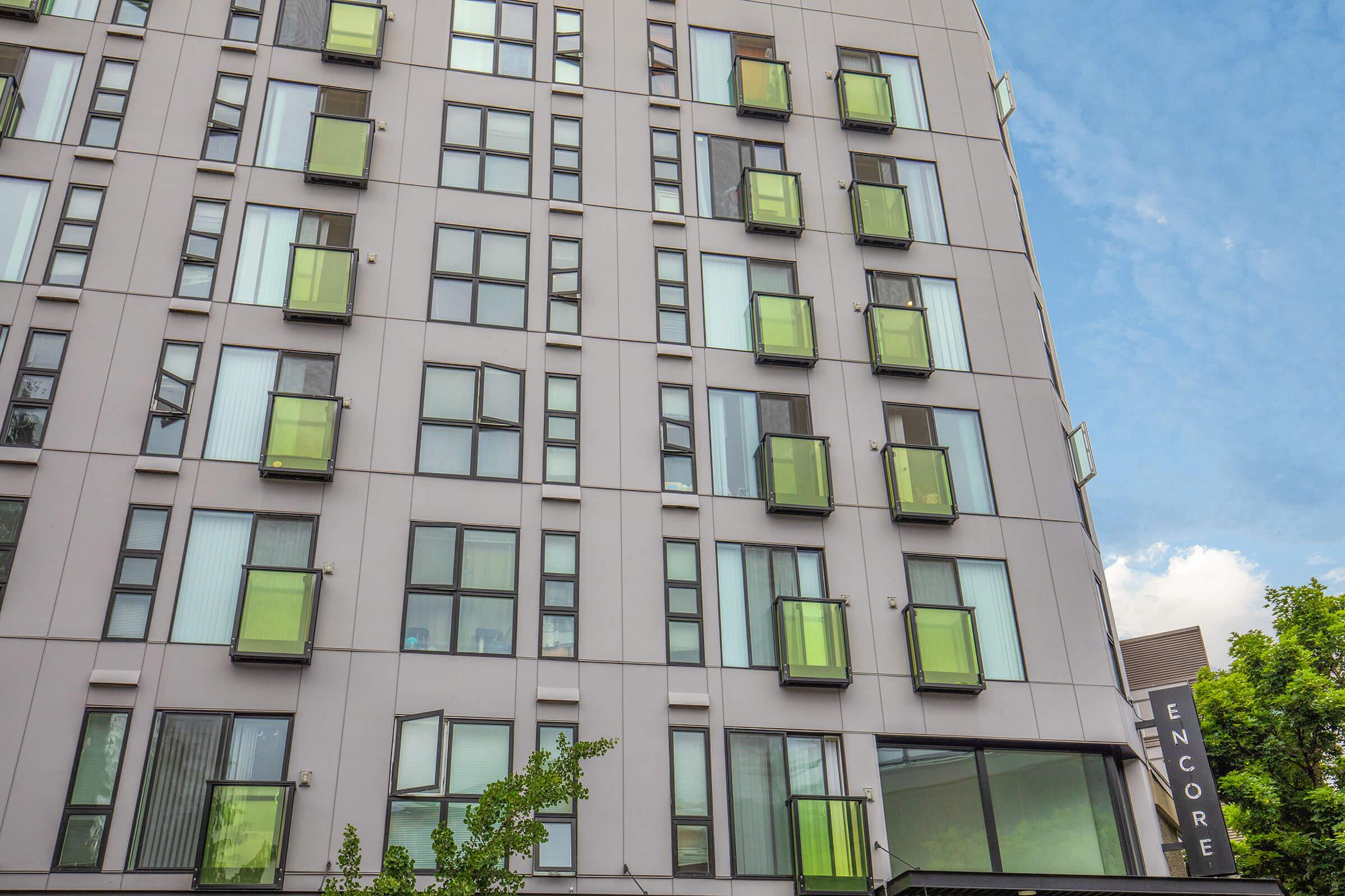 A modern apartment building with a gray facade featuring numerous square windows and distinctive green balconies. The sky is blue with some clouds, and part of a sign labeled "ENCORE" is visible on the right side of the image. Trees are partially in view at the bottom.