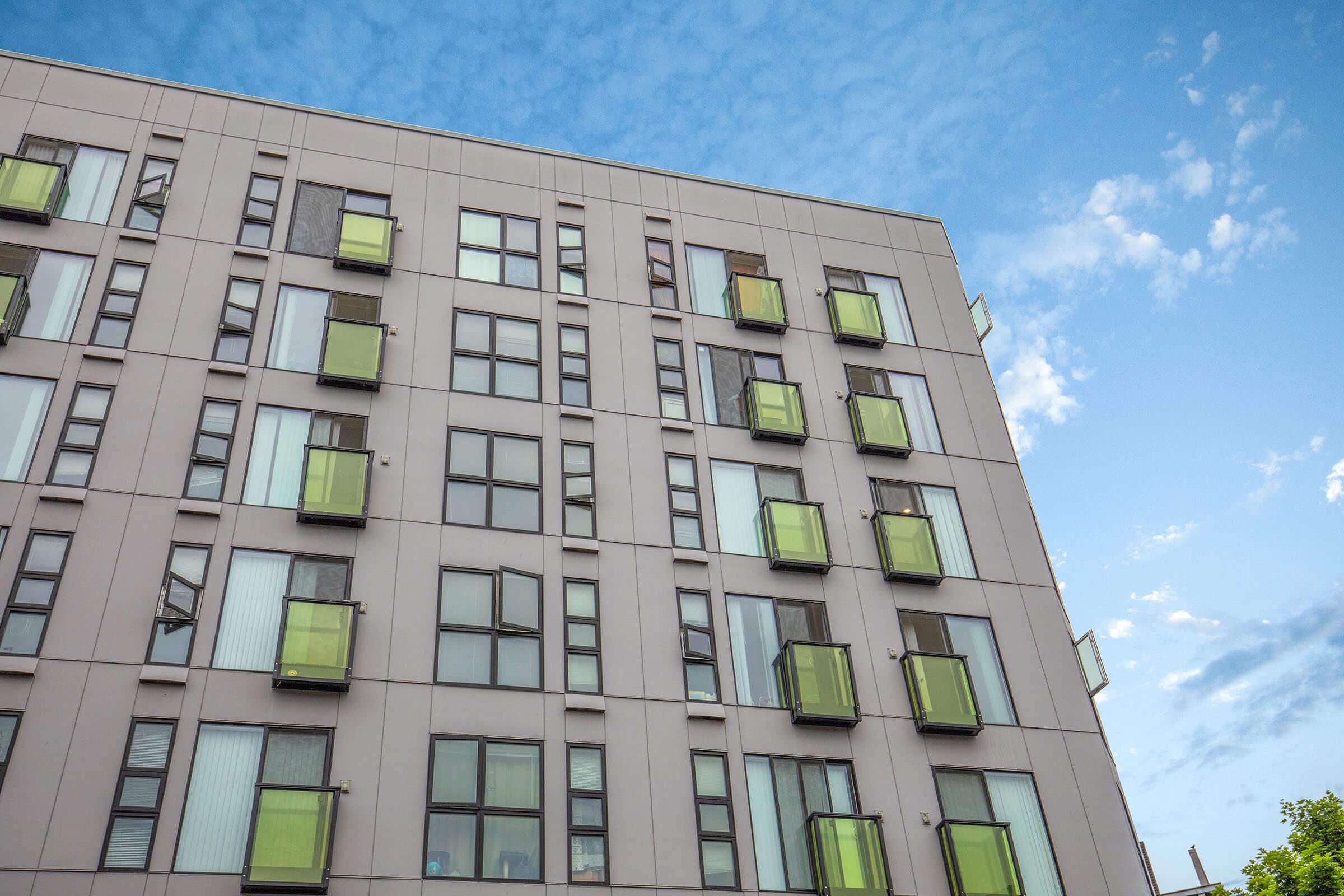 A modern apartment building with gray exterior walls and multiple large windows. Each window features a small green balcony. The sky above is partly cloudy with a blue background, enhancing the contemporary look of the architecture.