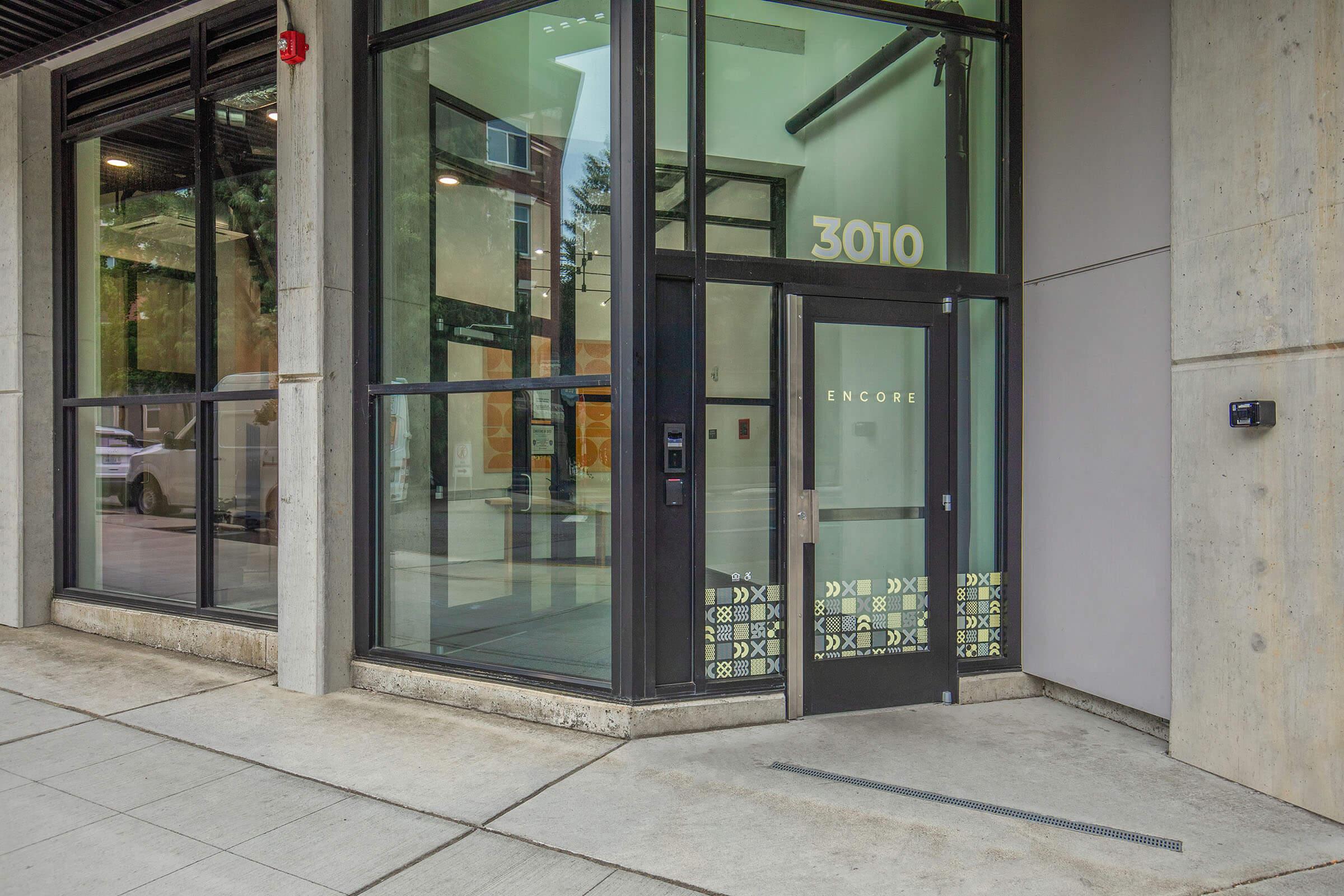 Entrance of a modern building featuring large glass windows, a sleek black door with the number "3010" displayed above. The door has decorative patterns at the bottom and the word "ENCORE" is visible. Concrete walls and a paved sidewalk are in the foreground.