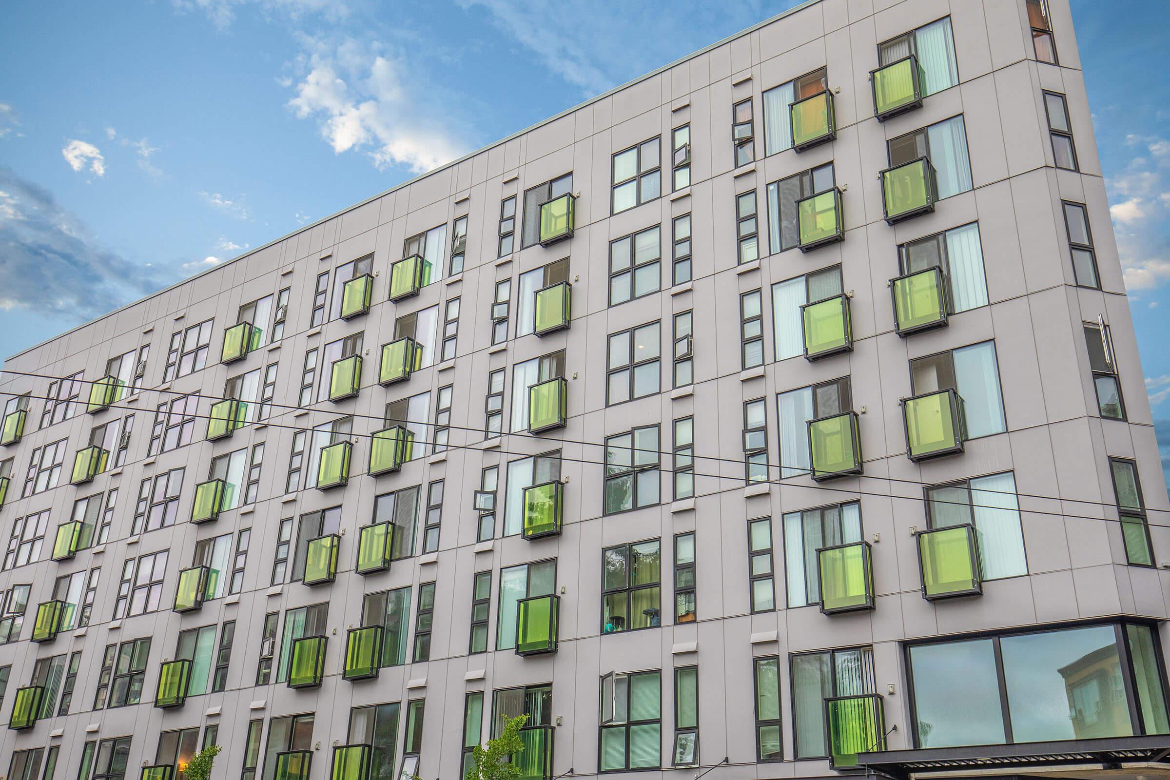 A modern building with multiple floors featuring large windows and uniquely designed green balconies. The structure has a sleek, contemporary facade, set against a blue sky with scattered clouds.