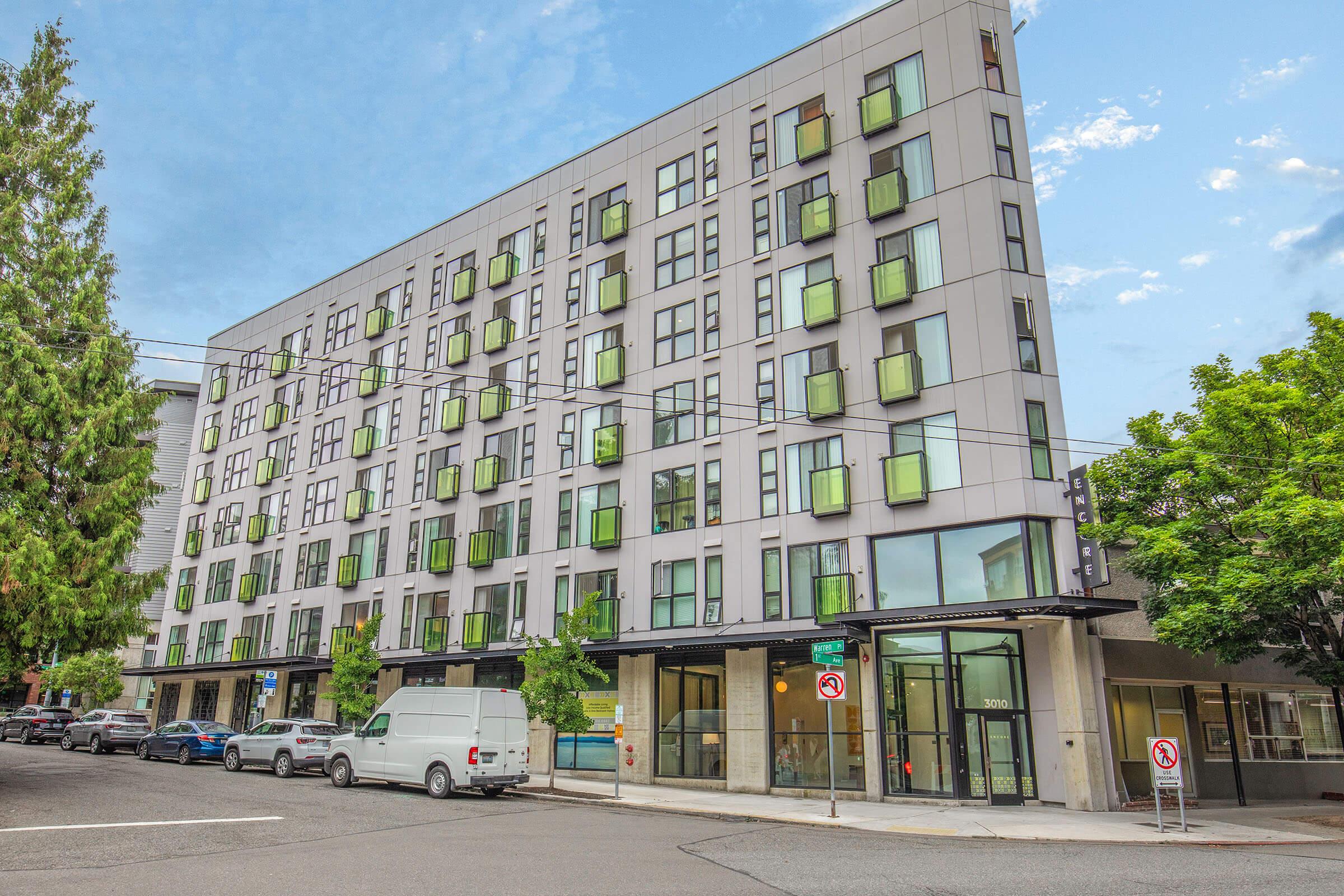 Modern multi-story building with a sleek gray facade and numerous large windows, reflecting green and blue hues. The entrance features glass doors, and the surrounding area includes parked vehicles and street trees under a partly cloudy sky.