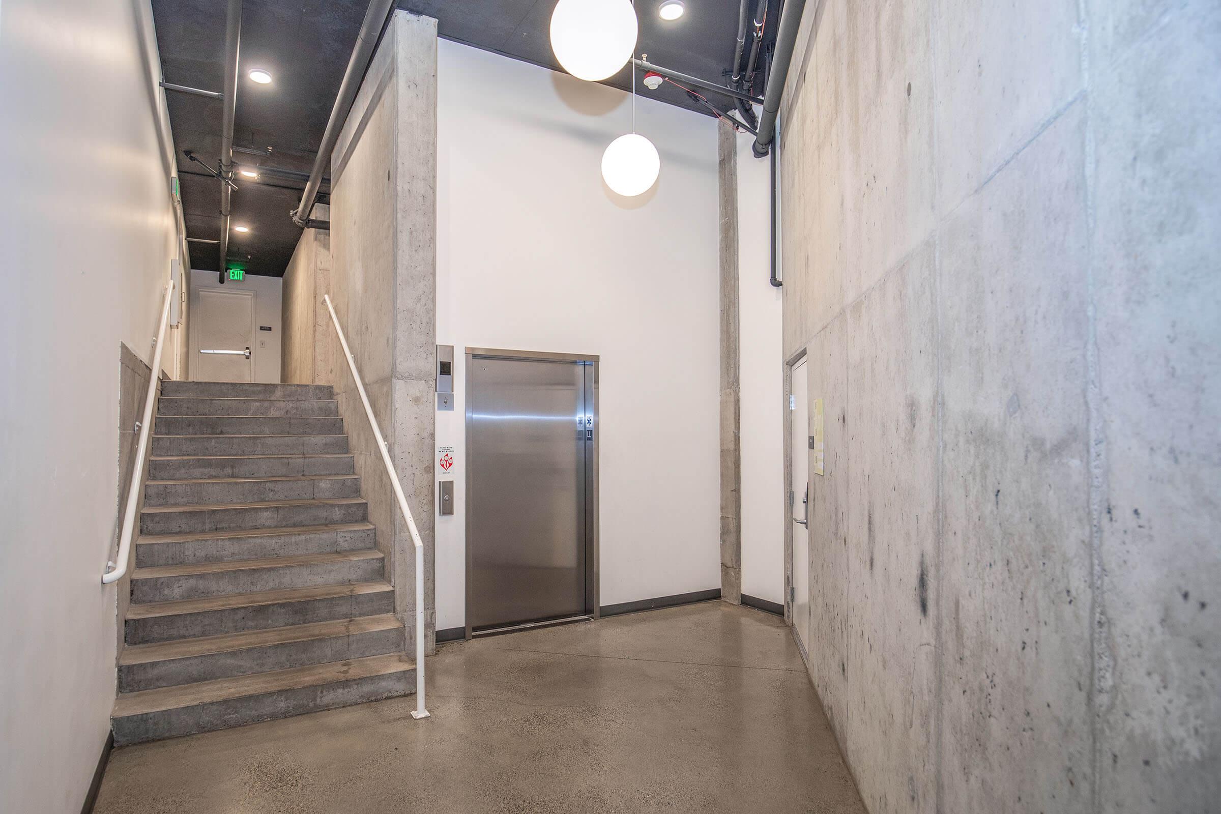 Stark corridor featuring a concrete wall and floor, with a staircase on the left and an elevator nearby. Two large pendant lights hang from the ceiling, illuminating the modern space. A door can be seen at the end of the hallway, adding to the minimalist aesthetic.