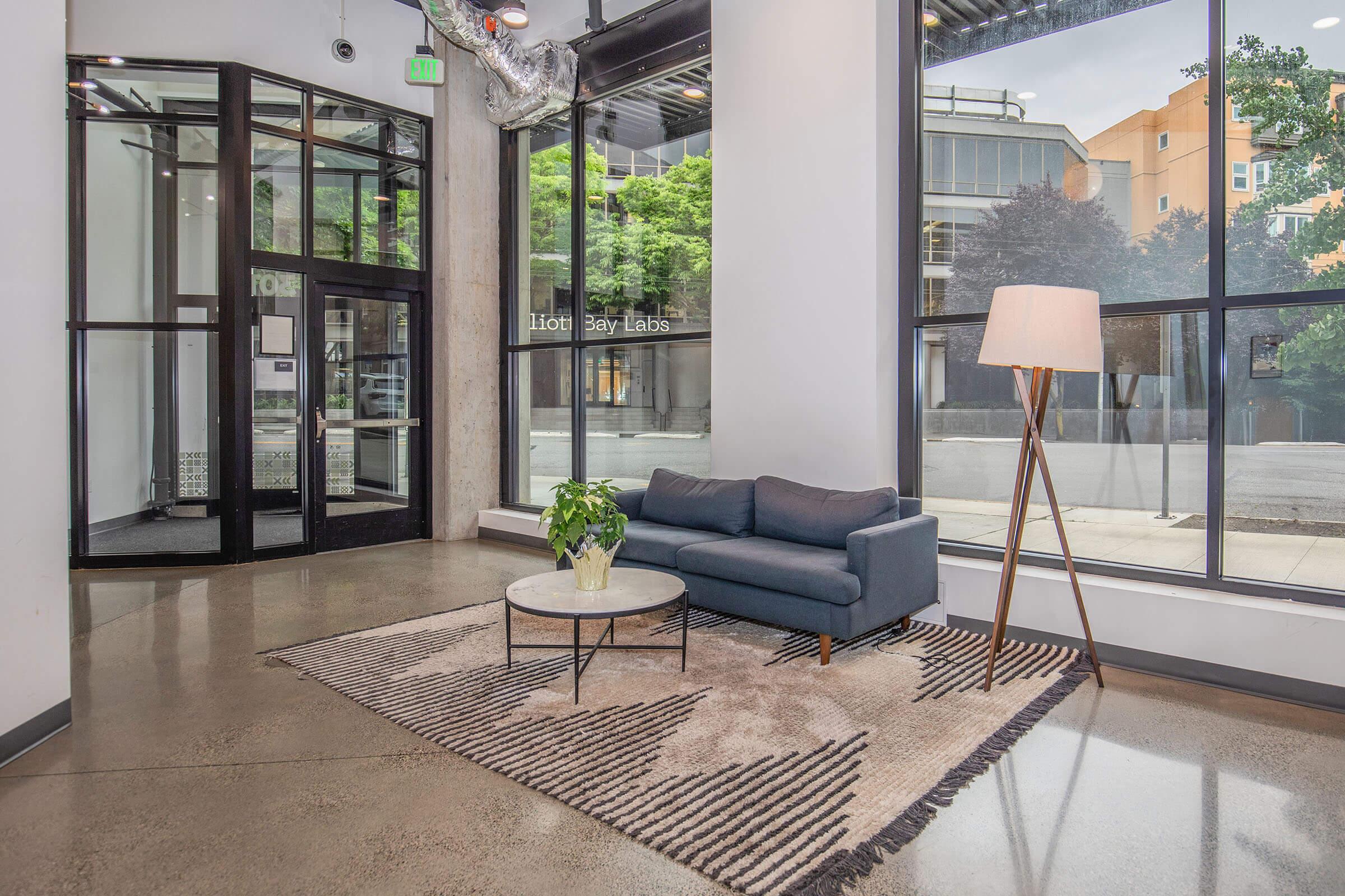 A modern interior lobby featuring large windows with green trees outside. There is a glass door entrance, a stylish gray couch placed on a patterned rug, a round coffee table, and a floor lamp, creating a welcoming and contemporary atmosphere.