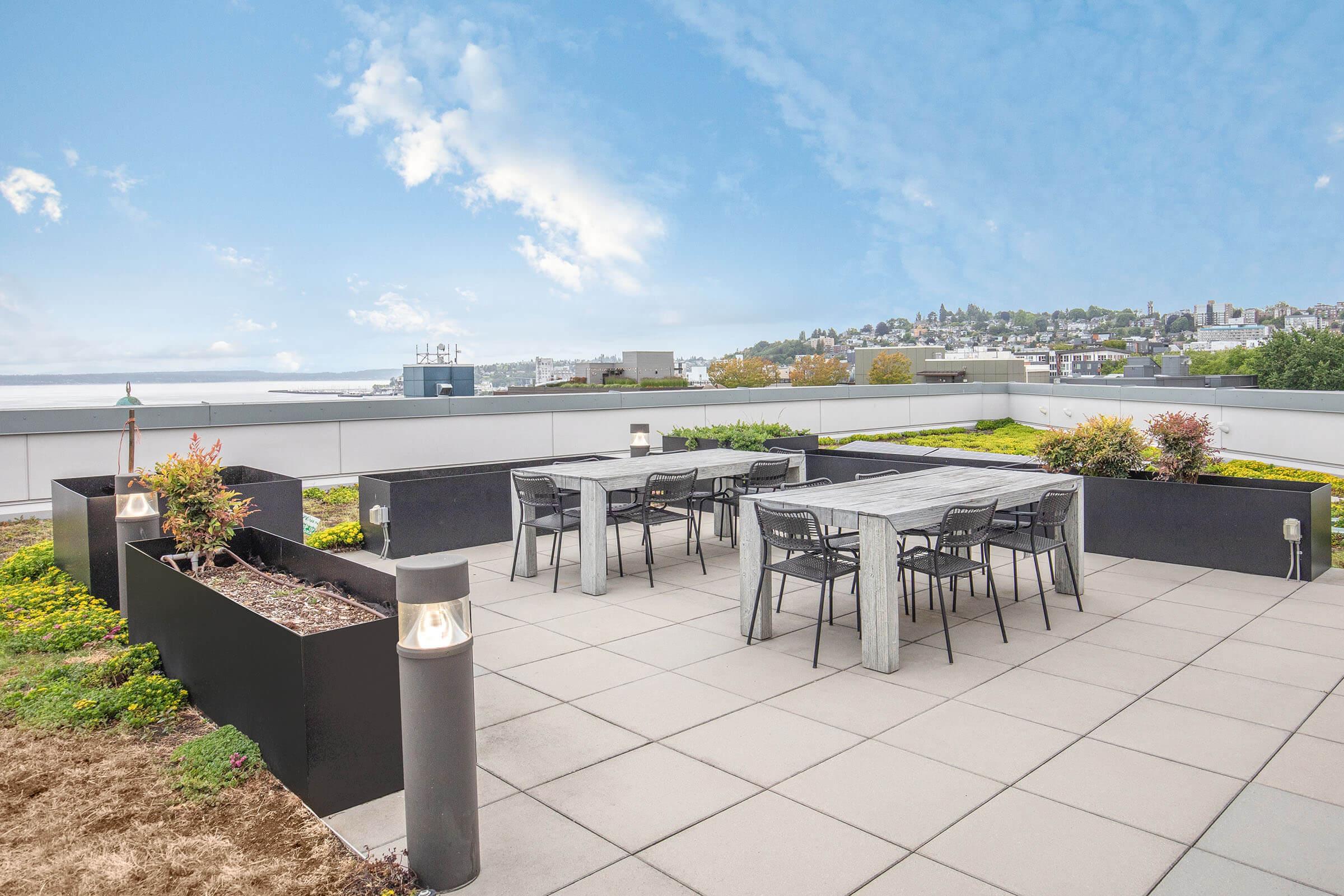 Rooftop terrace featuring a modern dining area with large tables and black chairs, surrounded by planters with greenery. A skyline view is visible in the background under a partly cloudy blue sky.