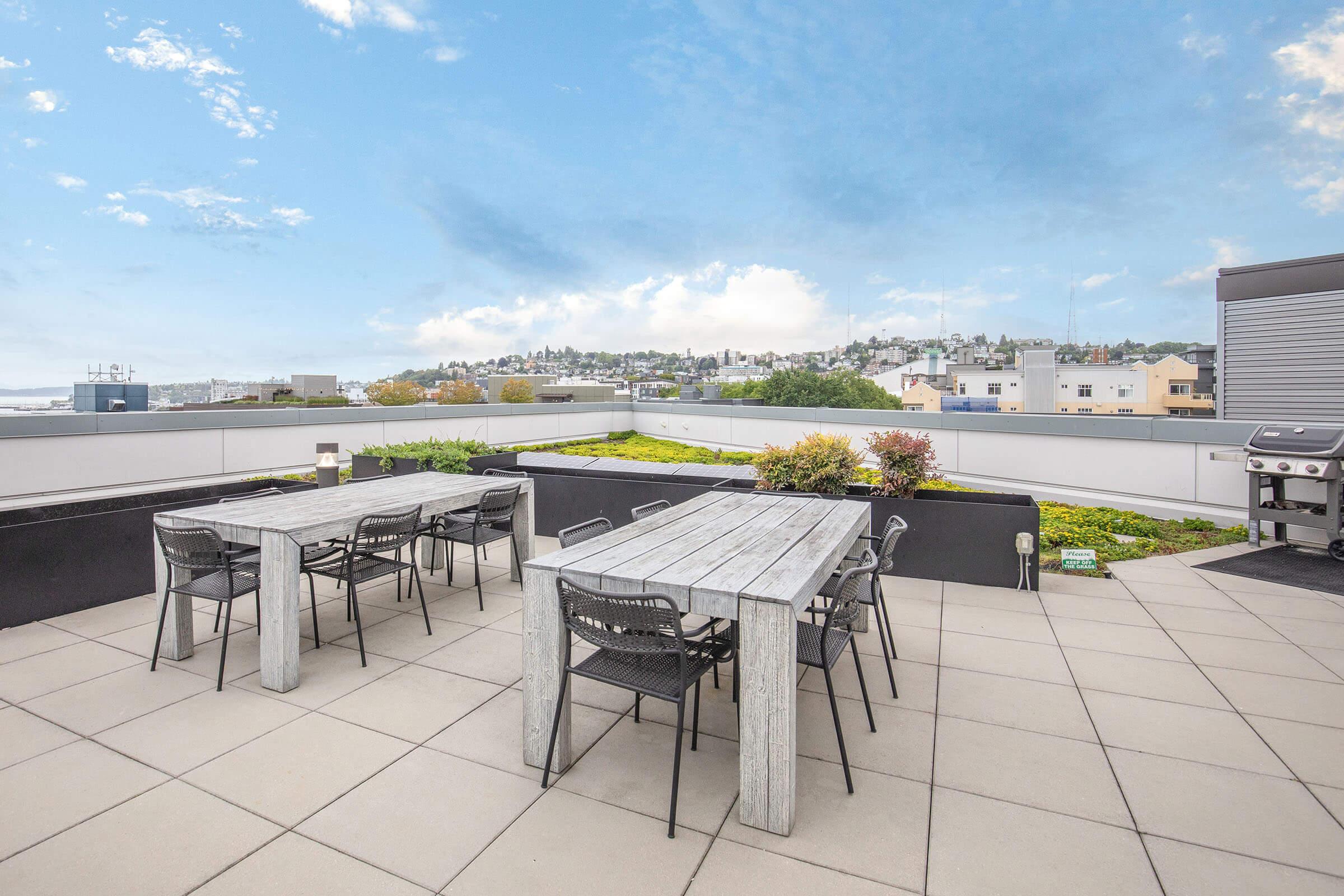 A modern rooftop terrace featuring multiple wooden tables with black chairs, surrounded by green plants. The area has a clear view of a city skyline under a bright blue sky with scattered clouds, and includes a barbecue grill on one side.