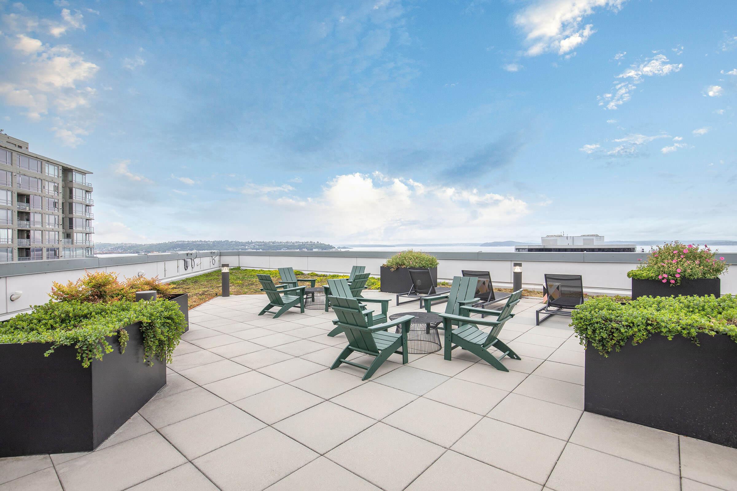 A rooftop terrace with green Adirondack chairs and a circular seating area surrounded by flower pots. The background features a tranquil view of the water and a partly cloudy sky, with a modern building in the foreground. The space is inviting for relaxation and gatherings.