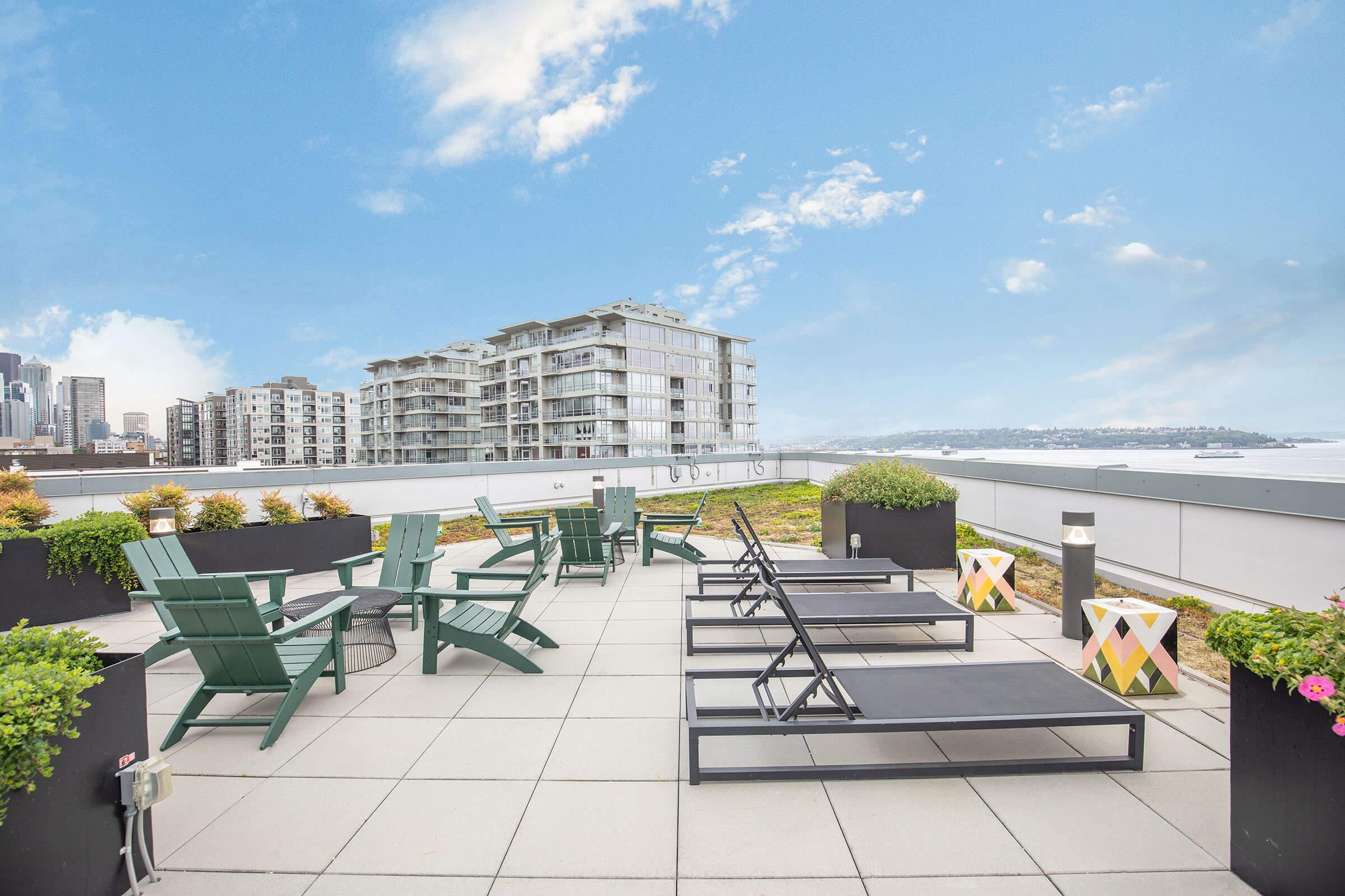 Rooftop terrace with green lounge chairs and sun loungers, surrounded by planters with greenery. A city skyline and waterfront are visible in the background under a clear blue sky. Ideal for relaxation or social gatherings.