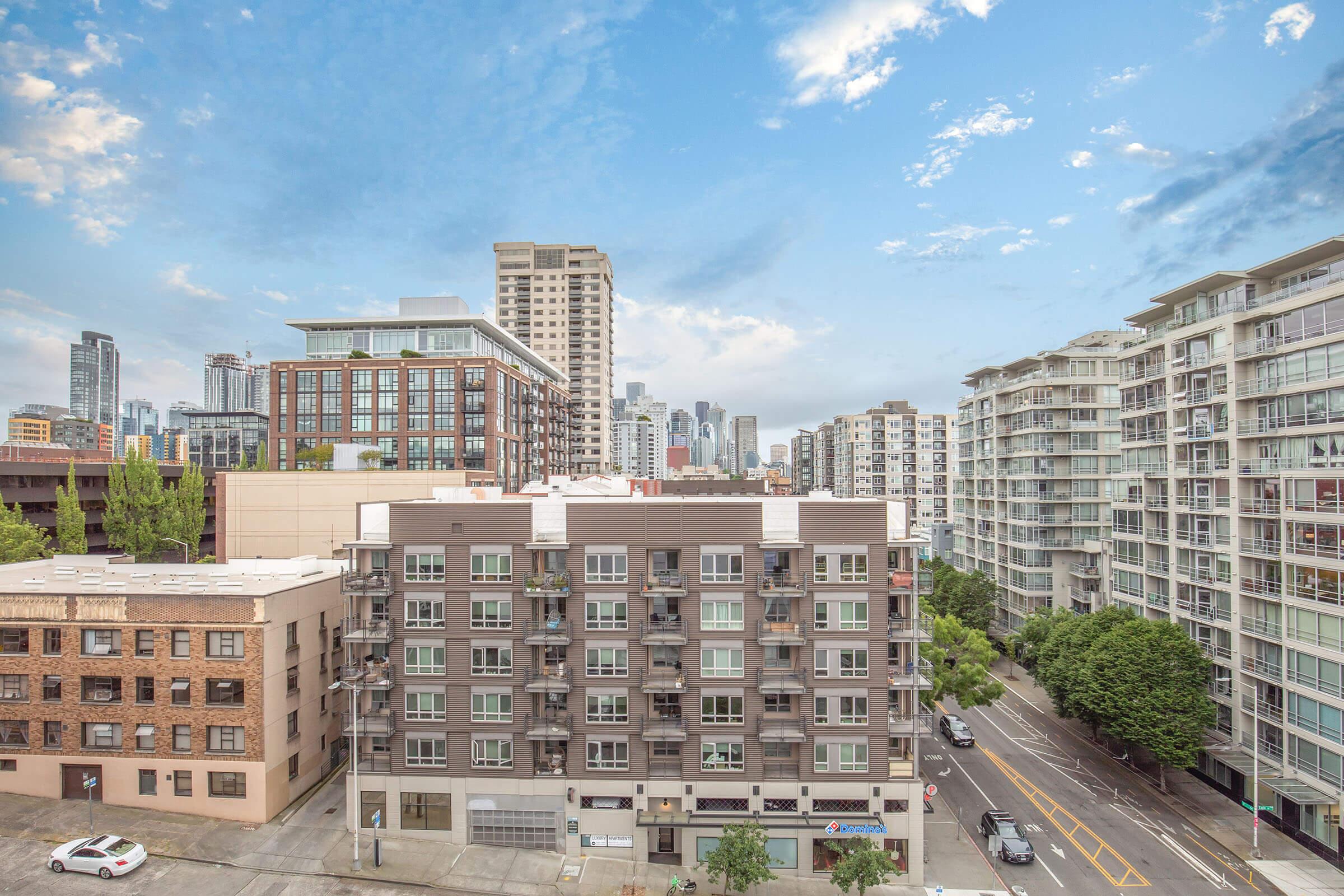 Cityscape view featuring a mix of modern and traditional buildings, with a skyline of high-rise structures in the background. The scene includes trees along the street and a blue sky with scattered clouds, capturing an urban environment.
