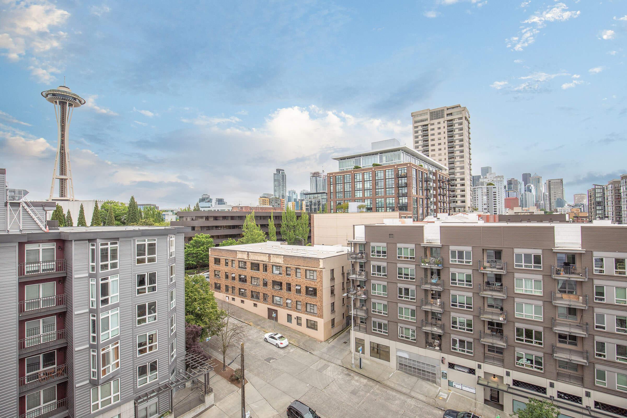 A cityscape view featuring a mix of modern and older buildings, with the iconic Space Needle in the background. The scene is set against a partly cloudy sky, showcasing a vibrant urban environment in Seattle. The image captures a blend of residential and commercial architecture.