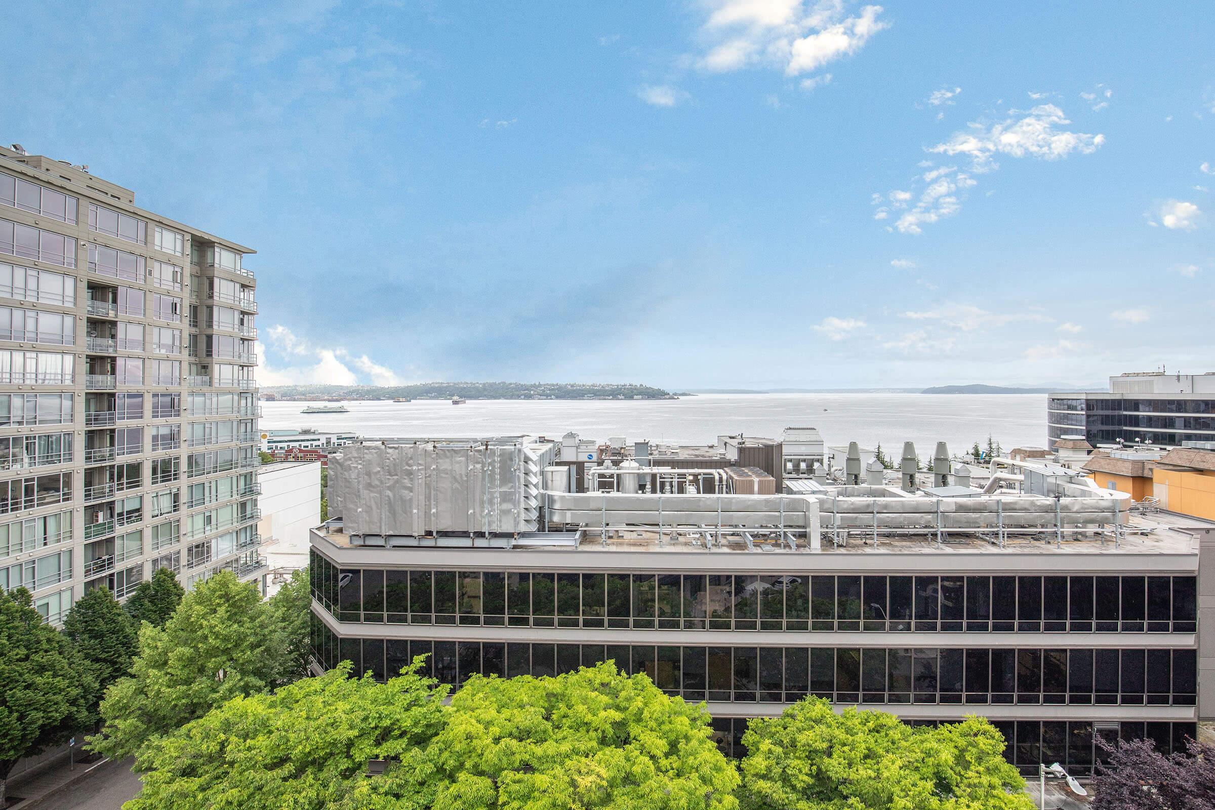 A panoramic view of a waterfront cityscape featuring modern buildings, green trees, and a calm body of water under a partly cloudy sky. The scene captures a blend of urban architecture and natural surroundings, highlighting a serene coastal atmosphere.