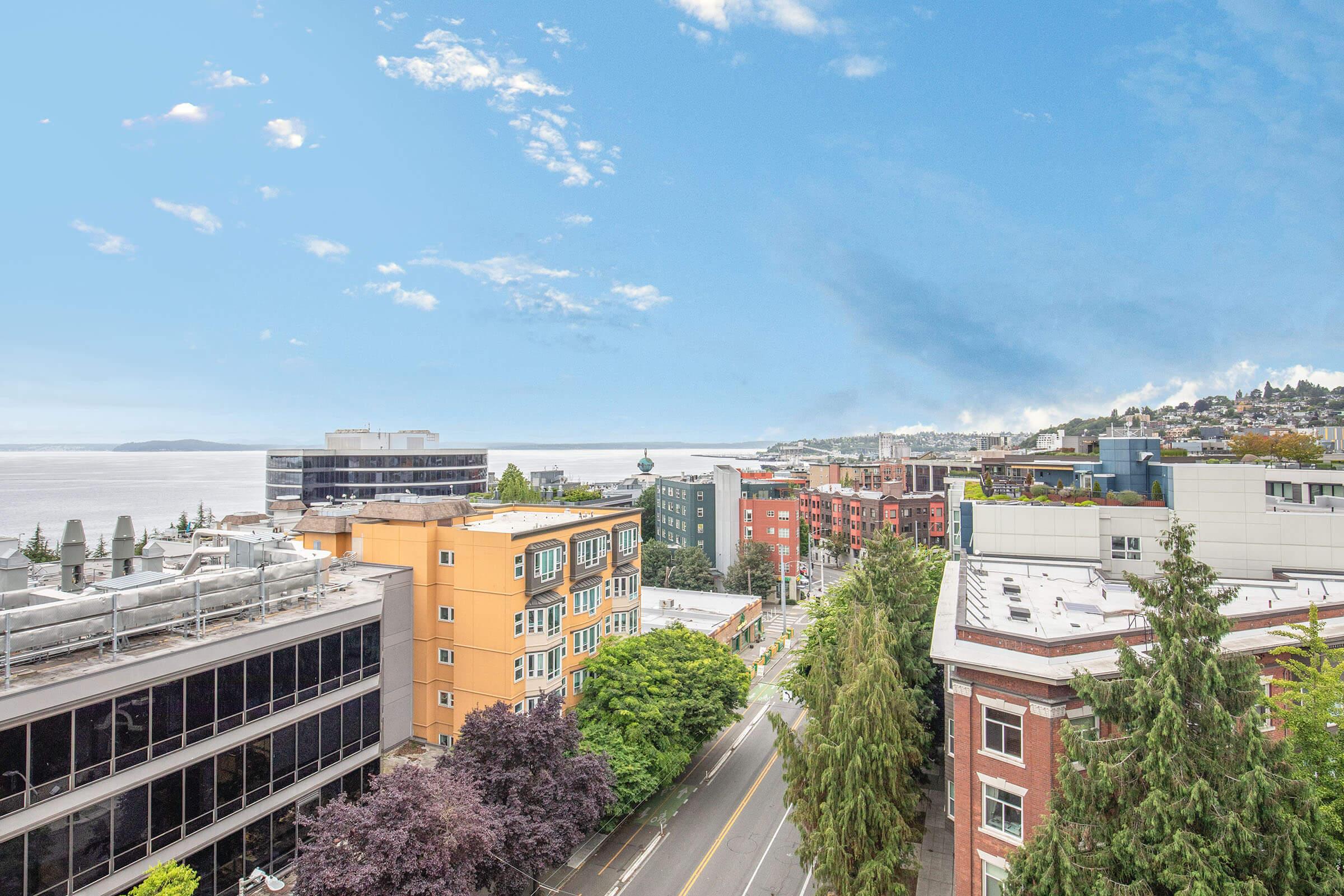 A panoramic view of a vibrant cityscape overlooking the water, featuring a mix of modern and historic buildings, lush green trees lining the street, and a clear blue sky with scattered clouds. The scene captures the essence of urban life alongside natural beauty.