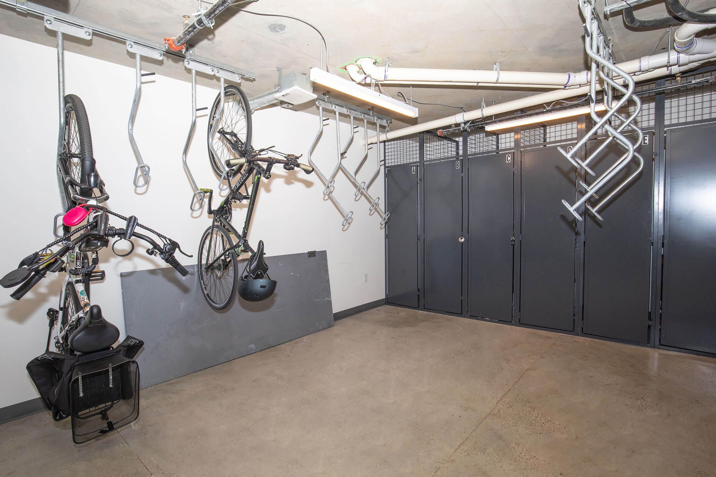 A clean bike storage area featuring two bikes hanging from the ceiling on hooks, with additional storage racks. The walls are bare, and there are several gray lockers on one side, providing secure storage options. The floor is concrete and the lighting is soft and functional.