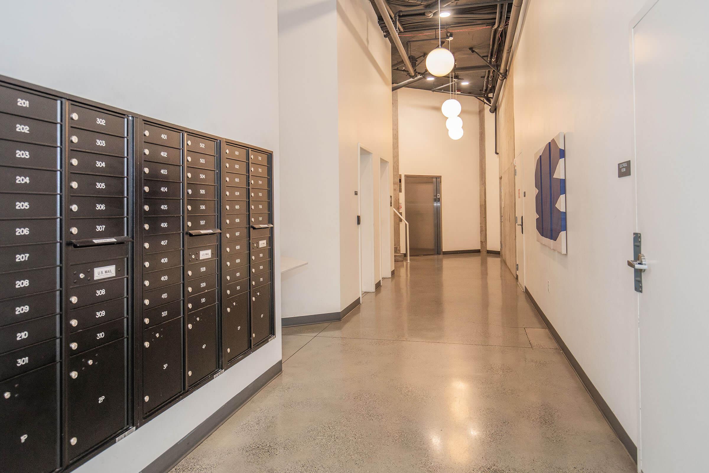 A modern hallway featuring a row of black mailboxes on the left side. The floor is polished concrete, and there are ceiling lights hanging above. An elevator door is visible at the end of the corridor, with abstract artwork hanging on the wall to the right. The space is well-lit and has a clean, minimalist design.
