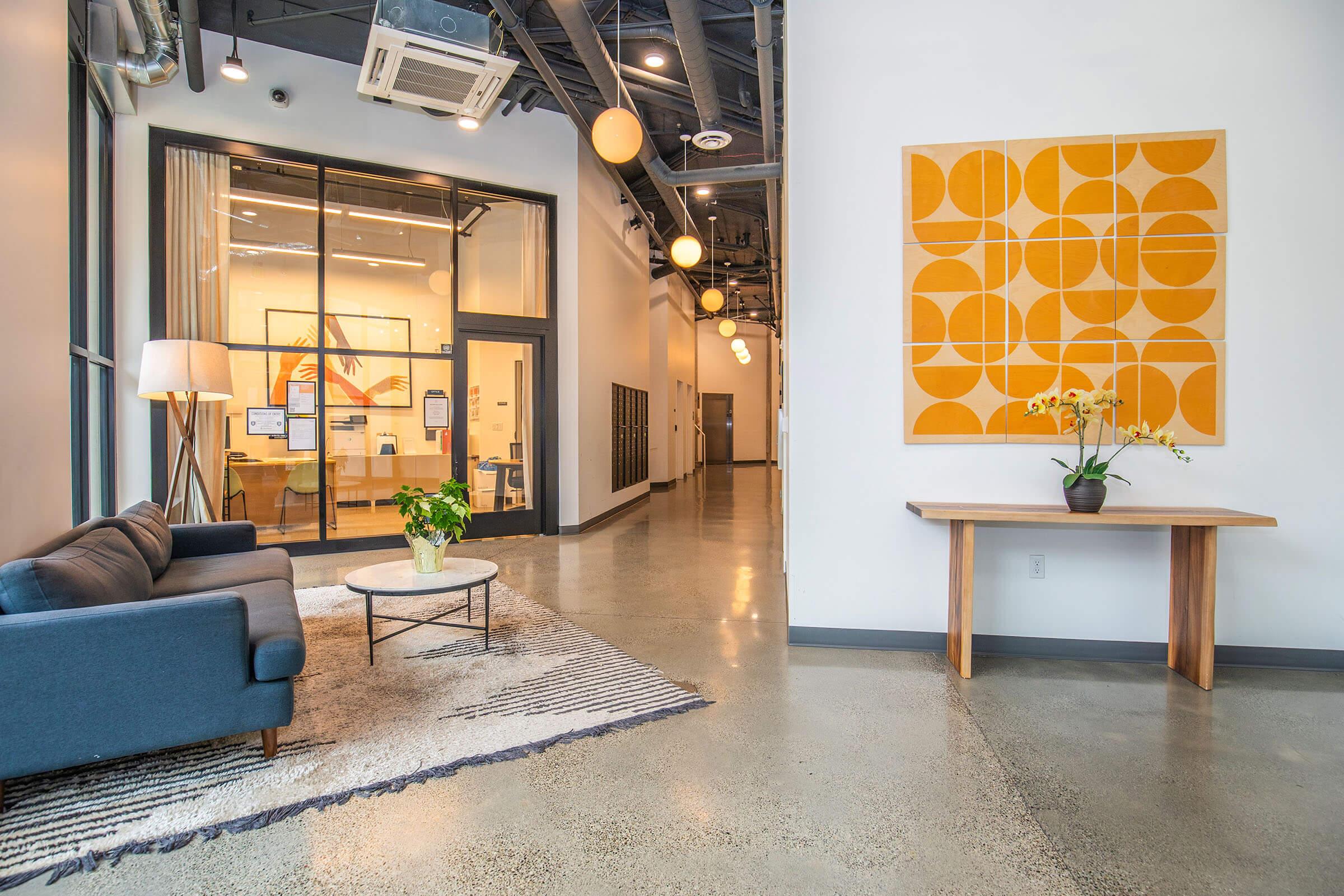 A modern, spacious lobby featuring a blue sofa and a circular coffee table on a patterned rug. A tall lamp stands beside the sofa. On the wall, a large abstract artwork with orange shapes is displayed. In the background, a glass office area is visible, along with a small potted plant on a wooden table.