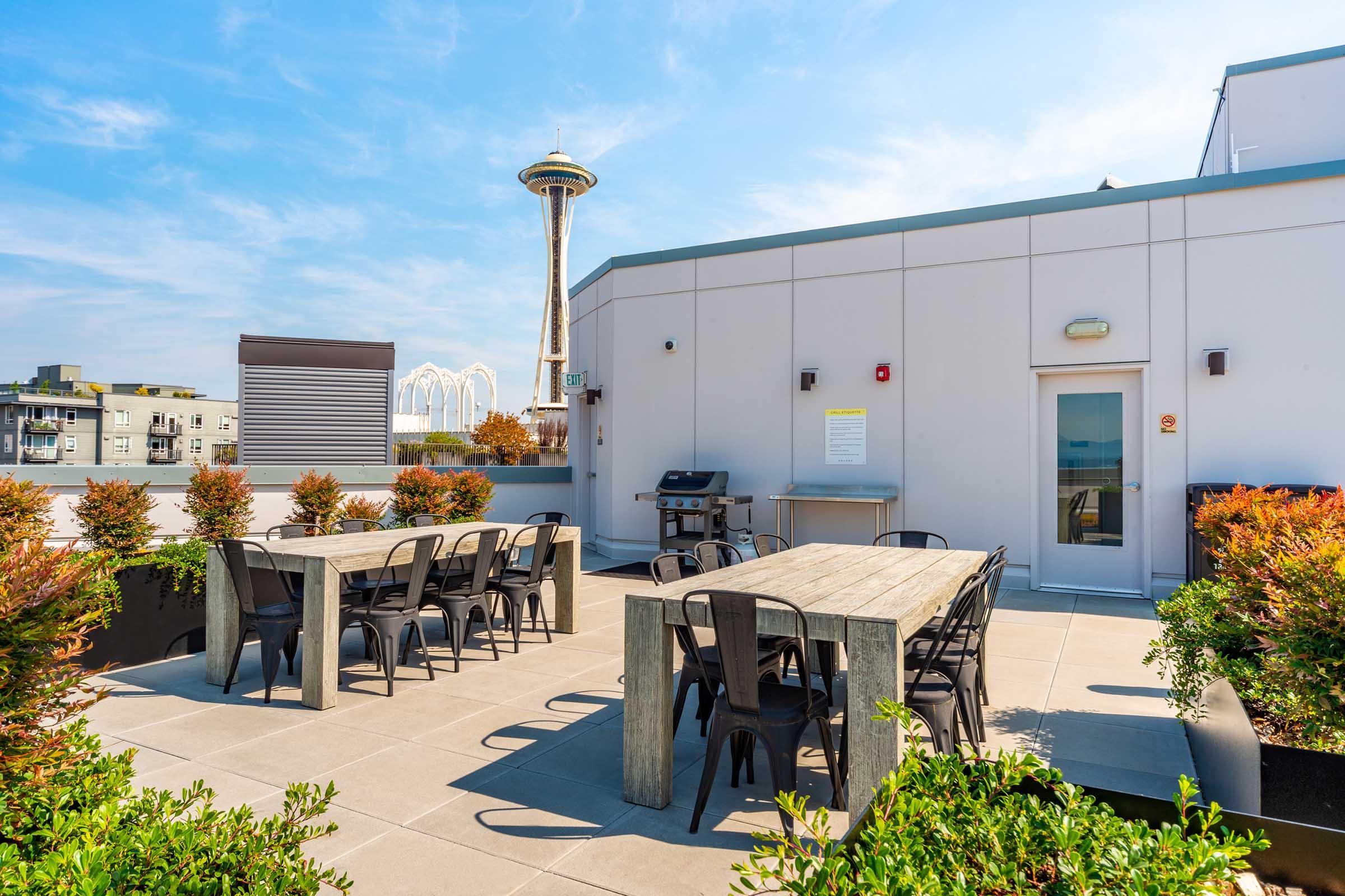 A rooftop terrace featuring modern outdoor dining tables and chairs surrounded by plants. The Space Needle is visible in the background under a bright blue sky. A grill and a door lead to an indoor area, creating an inviting space for gatherings and relaxation.