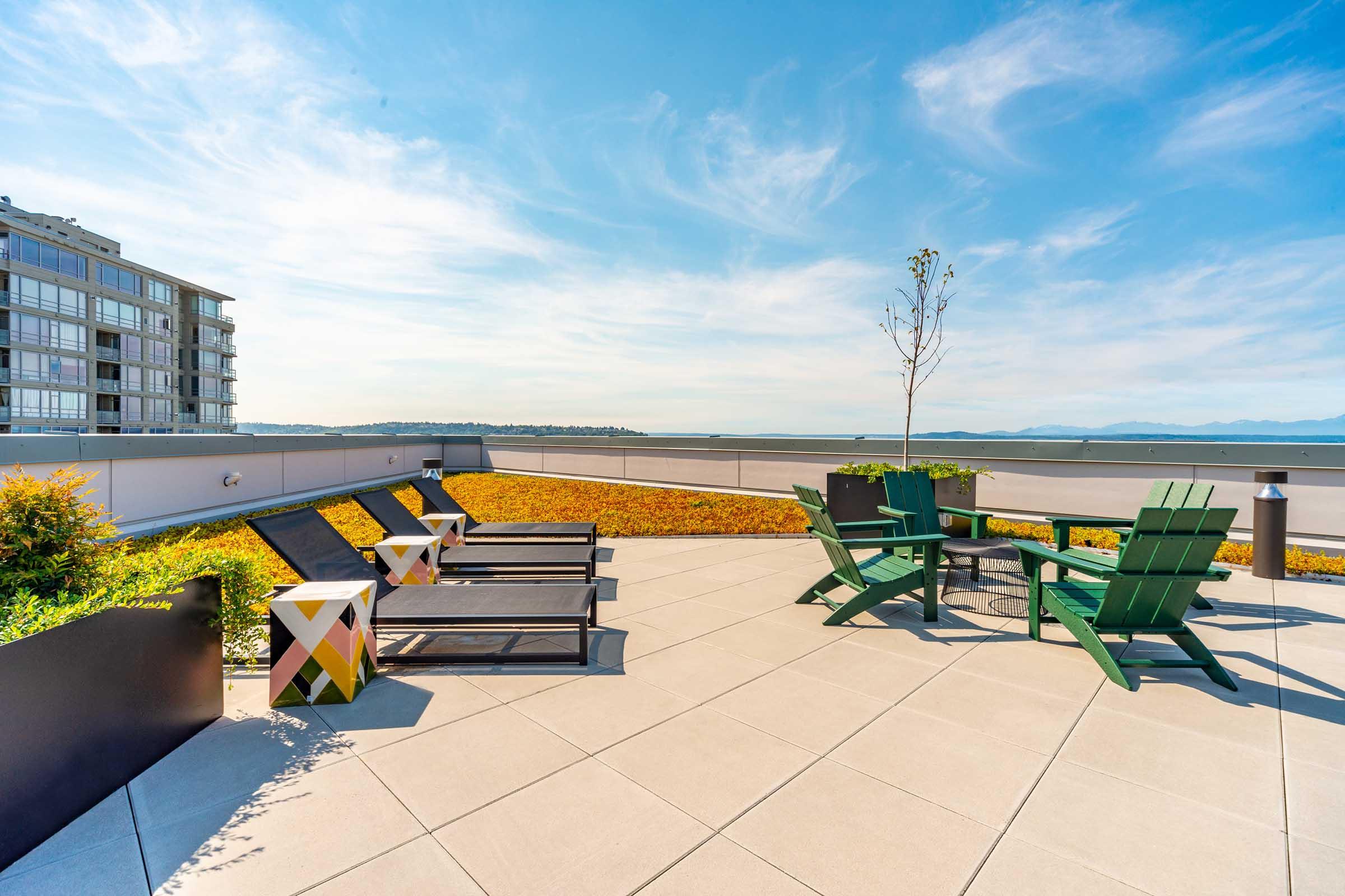 A modern rooftop terrace featuring green Adirondack chairs, lounge chairs, and a small table, surrounded by vibrant landscaping. The view includes water and mountains in the distance under a bright blue sky with wispy clouds. The setting is designed for relaxation and outdoor enjoyment.