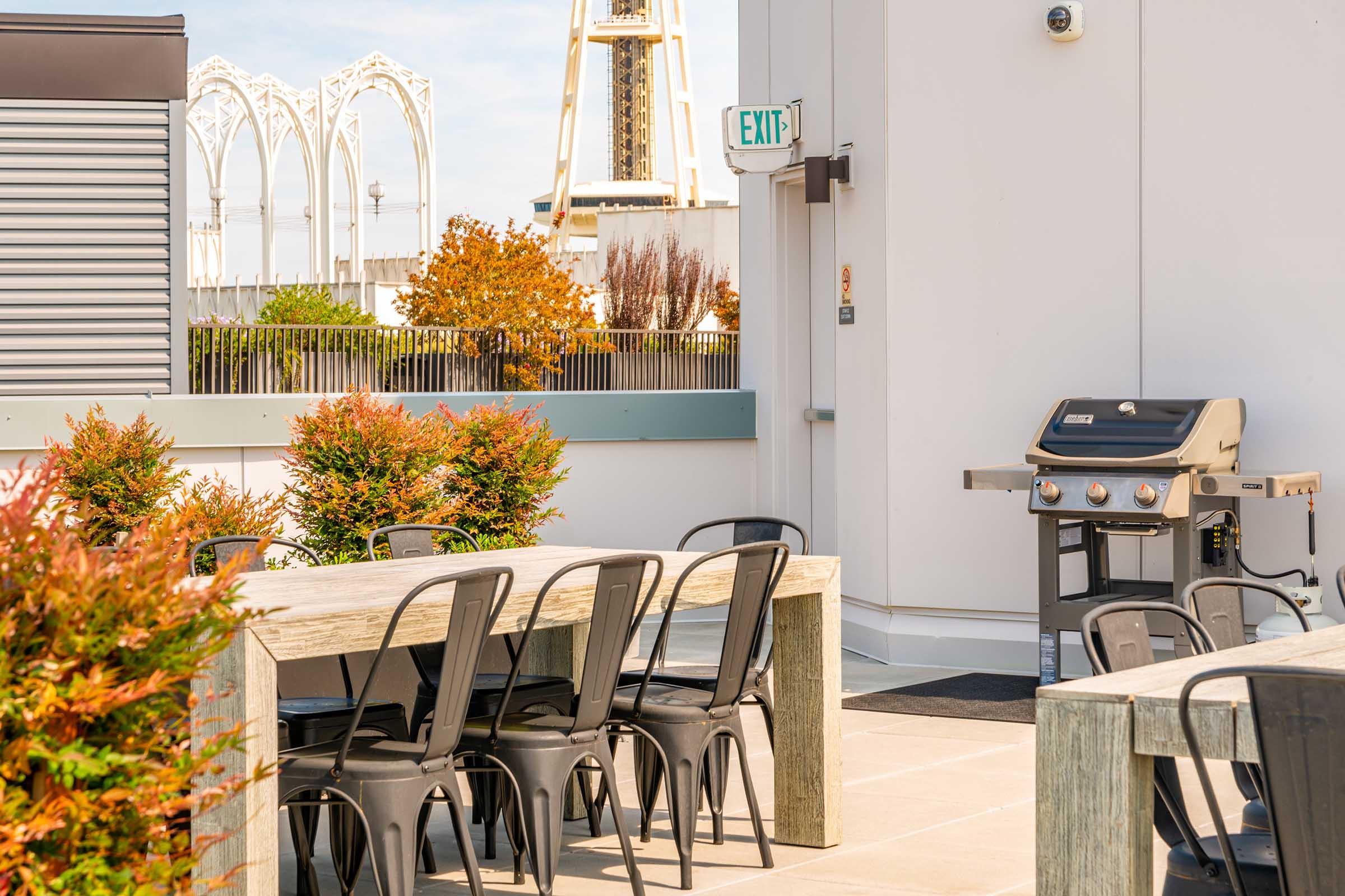 A rooftop terrace featuring a large wooden table surrounded by black metal chairs. A gas grill is positioned nearby, with colorful autumn foliage in the background and a partially visible structure resembling a ferris wheel against a clear sky. An exit sign is also visible.