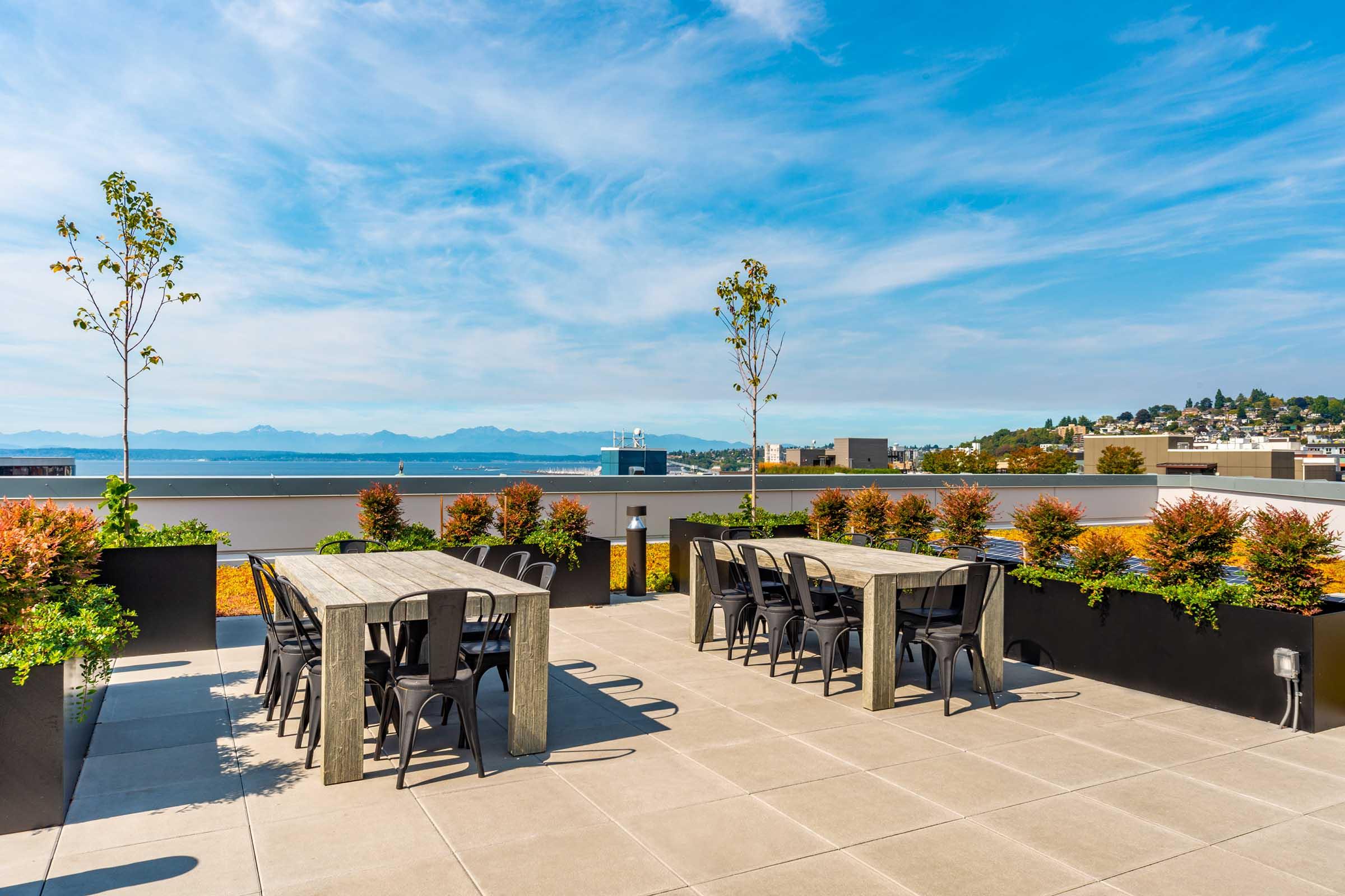 Rooftop terrace with several tables and chairs, surrounded by planters filled with colorful plants. In the background, a scenic view of distant mountains and a body of water under a clear blue sky with scattered clouds.