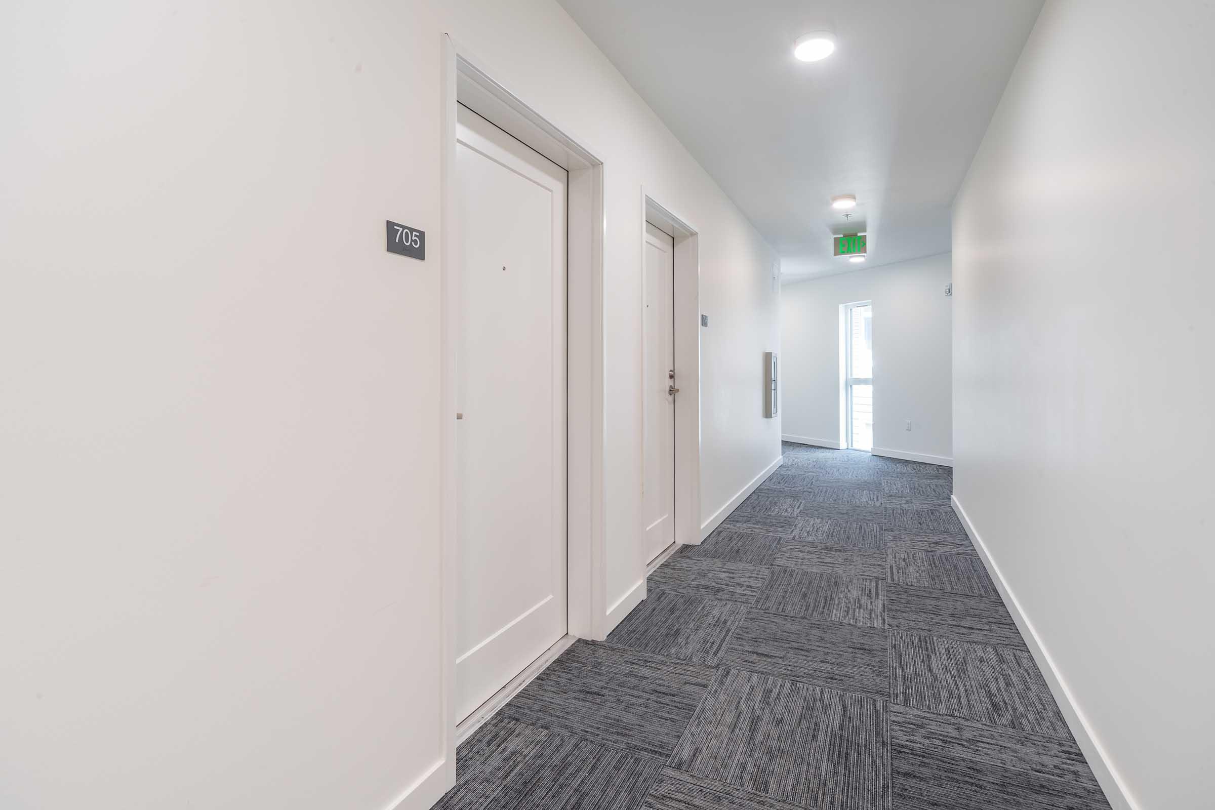 A well-lit, modern hallway featuring two closed doors on the left marked with room numbers. The walls are painted white, and the floor is covered with dark carpet tiles. A window at the far end allows natural light, creating a spacious and inviting atmosphere.