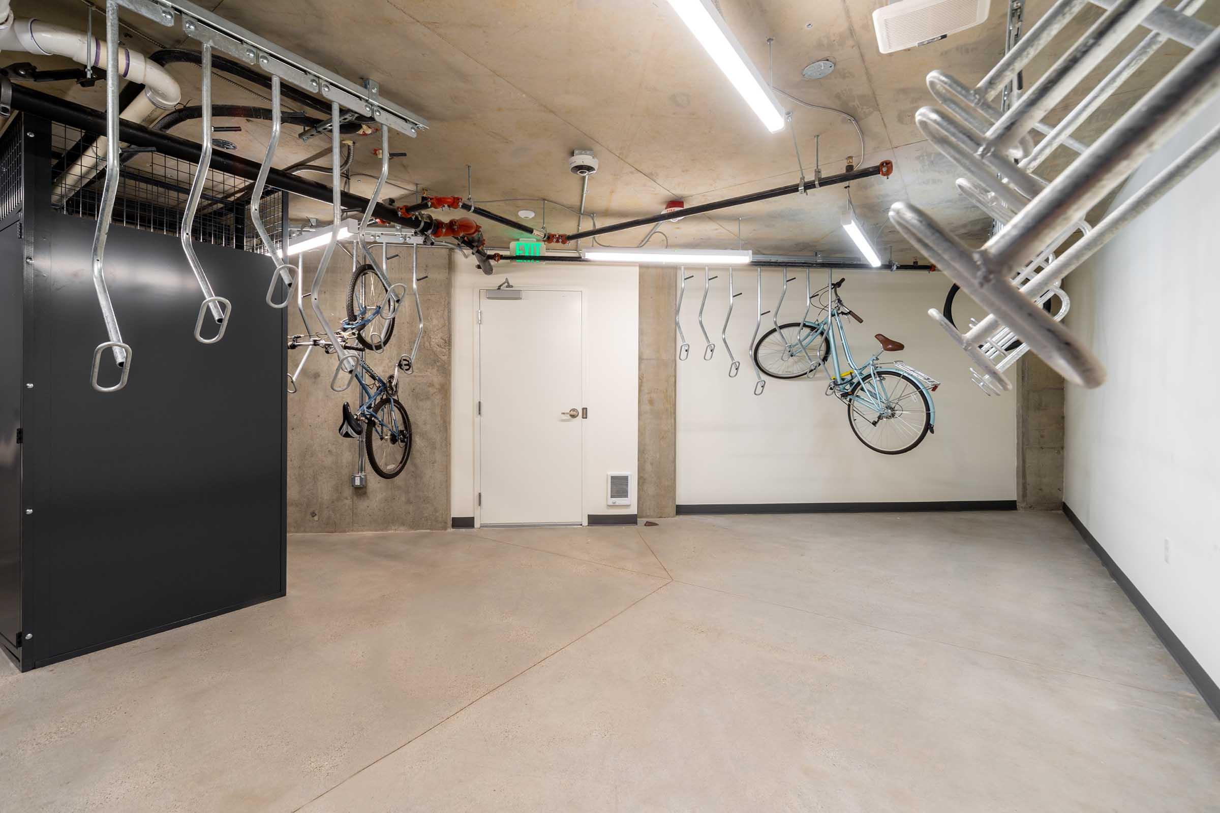 A spacious indoor bike storage area with grey concrete walls and flooring. Several bicycles are mounted on hooks hanging from the ceiling, and a black storage unit is in the corner. A white door is visible on one wall, providing access to the space. Overhead lighting brightens the area.