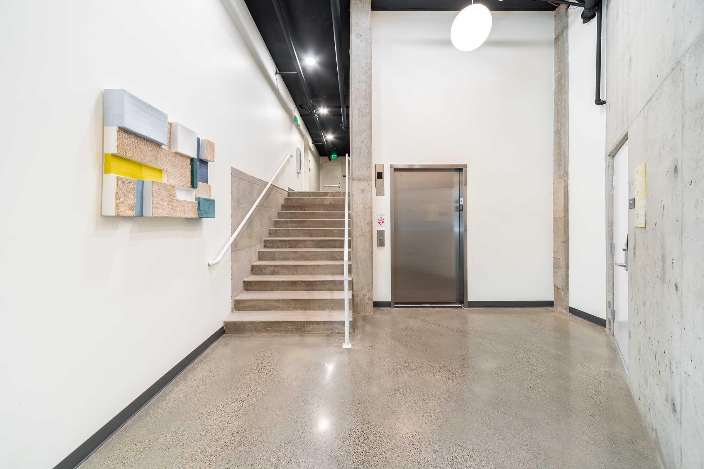 A well-lit hallway features a textured wall hanging on the left, leading to a staircase on the right. An elevator is situated next to the stairs, with polished concrete floors reflecting the overhead lighting. The walls are painted in neutral tones, creating a modern and inviting atmosphere.