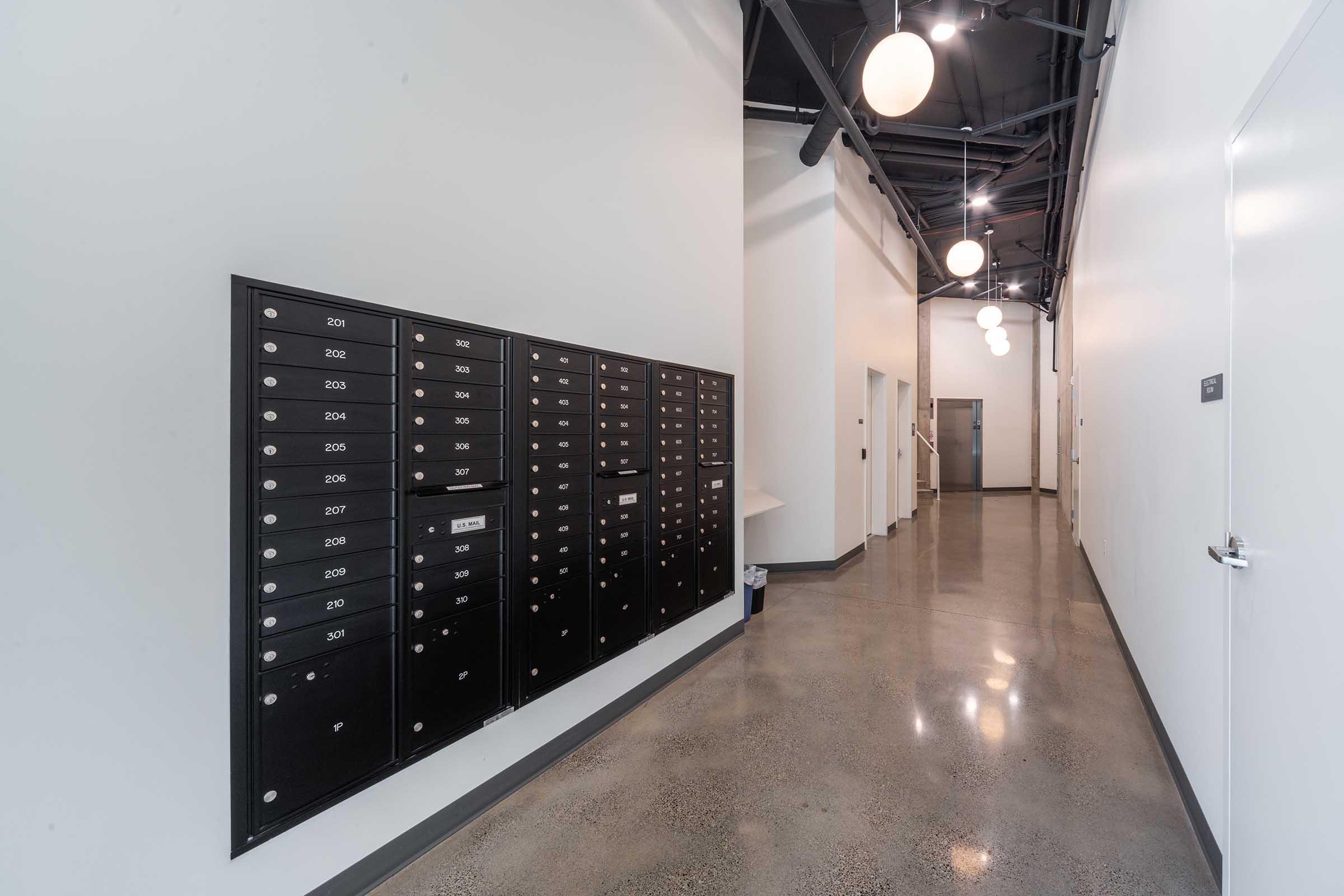 A hallway featuring a black mailbank with multiple locked mailboxes on the left side. The space has concrete flooring, white walls, and modern lighting fixtures hanging from the ceiling. Doors to rooms are visible in the background, creating a sleek, minimalist environment.