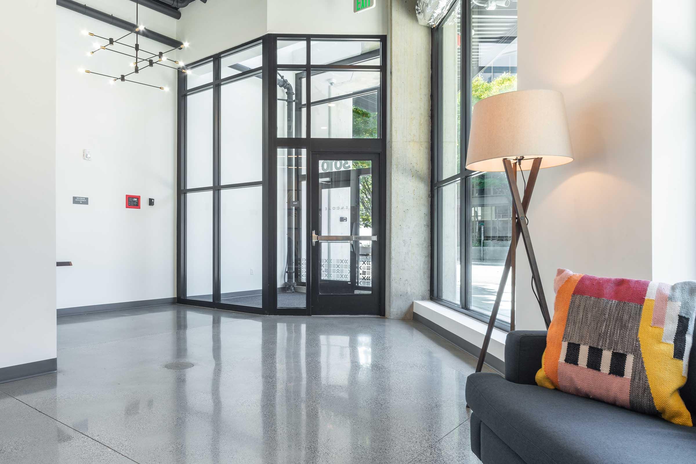 Interior of a modern lobby featuring a glass entrance with a large door, polished concrete flooring, a stylish floor lamp, and a couch adorned with a colorful geometric pillow. The space is bright with natural light coming through large windows.