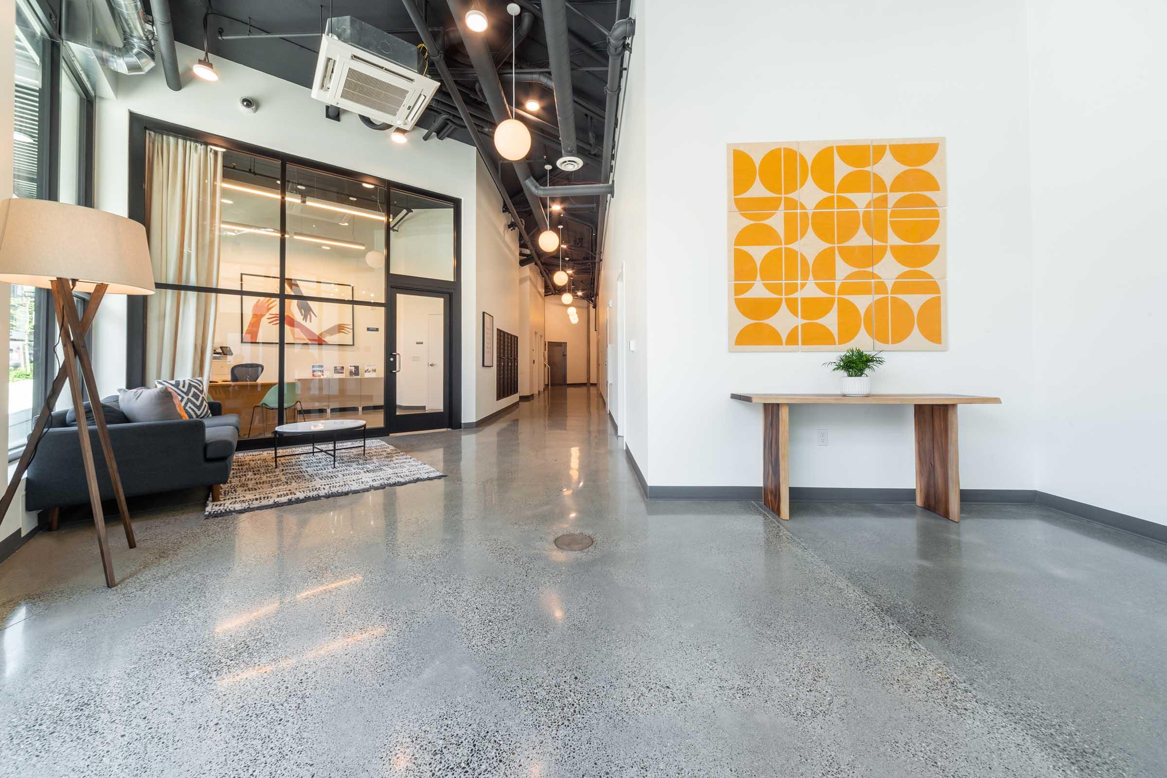 Modern interior featuring a spacious hallway with polished concrete floors, a contemporary table, a stylish lamp, and a decorative orange wall art piece. Natural light pours in through large windows, and the space is accented with minimalist furniture and decor, creating an inviting atmosphere.