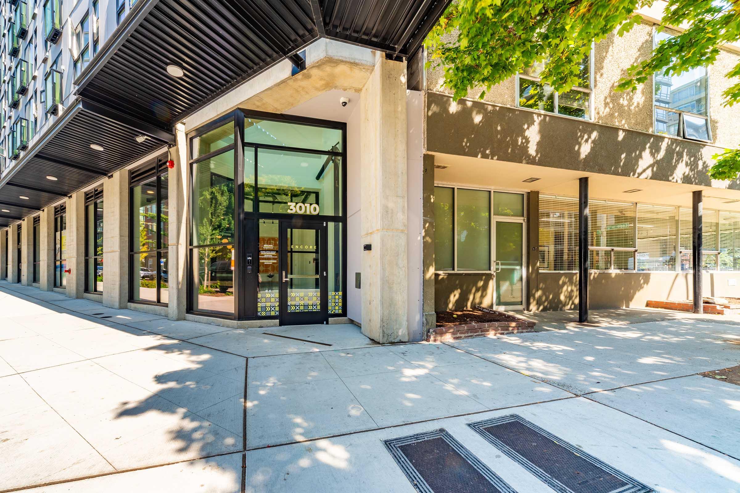 A modern building entrance featuring large glass doors and a concrete facade. The corner of the building is highlighted by a row of windows, and lush green trees provide shade over the sidewalk. The address "3010" is prominently displayed on the door.