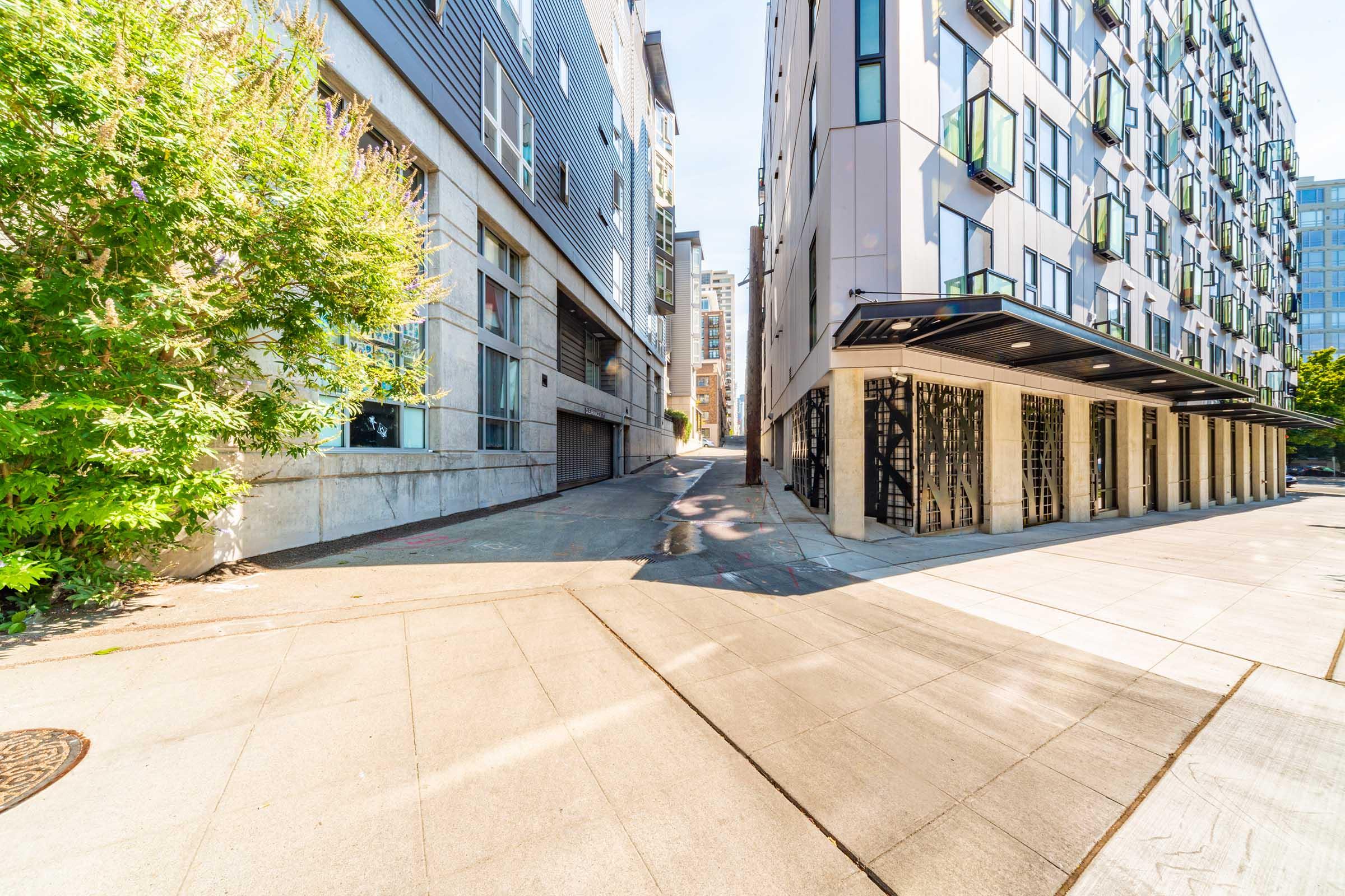 A streetscape featuring a modern urban environment. On the left, a building with a reflective glass facade and a textured concrete wall. On the right, a multi-story apartment building with balconies. The pathway between them slopes gently upward, surrounded by greenery and urban architecture under a clear blue sky.